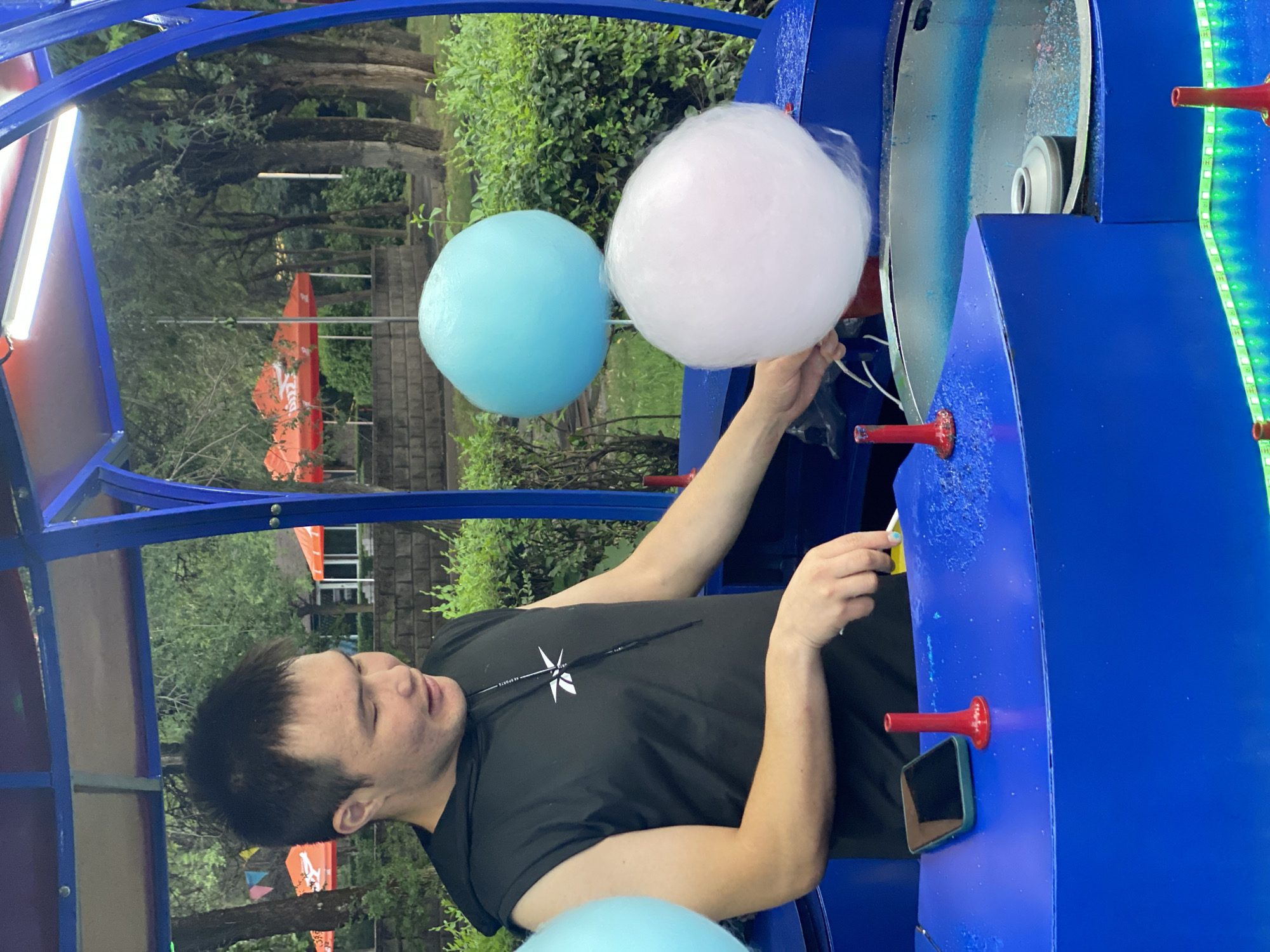 The image shows a young man making cotton candy at an outdoor amusement park in Almaty, Kazakhstan. He is wearing a black t-shirt and appears to be in the process of pulling a cone of pink cotton candy away from a spinning machine. To his left, a blue ball of cotton candy is also visible. The background features lush green trees and foliage, suggesting a pleasant day, possibly in the late afternoon or early evening, given the lighting. The cotton candy machine is blue and has red holders for the cones. The man's expression is focused on his task. The scene is set in an area with park-like landscaping, with hints of amusement park structures and decorations visible. There is a visible logo on the man's t-shirt which reads "AE SPORTS".