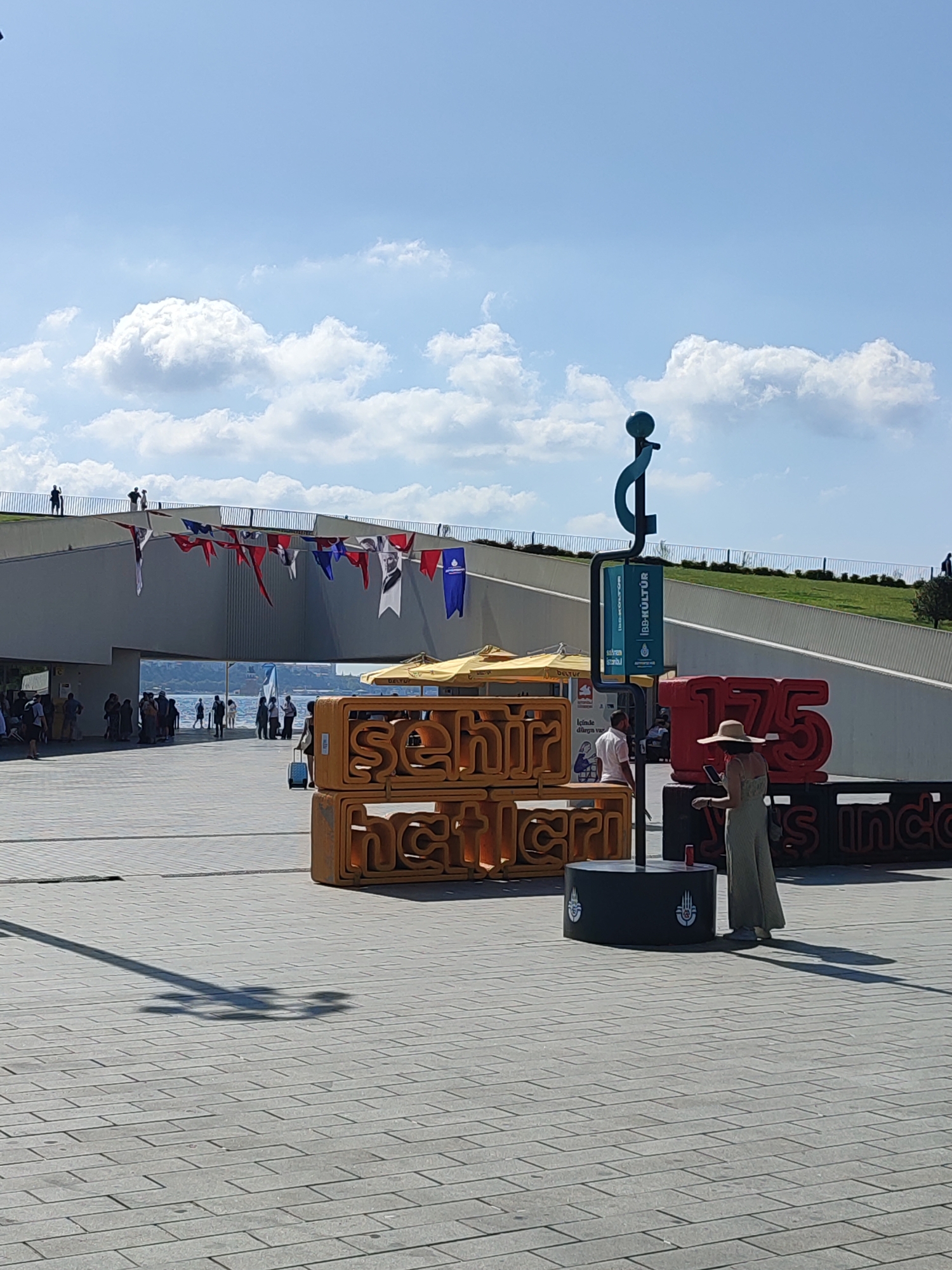 The image depicts an outdoor scene in the city of Beyoğlu, Turkey, under a bright, partly cloudy sky. In the foreground, a paved walkway is visible, with a large shadow of a stylized question mark-like sculpture cast upon it. To the right, a woman wearing a straw hat and a long, light-colored dress is standing and looking at her phone. She is positioned near a large, red, three-dimensional numeral "175" and a black cylindrical object with white emblems. Behind the woman, there are large, yellow, three-dimensional letters spelling out "sehir" above "netleri". Further back, a modern, white, elevated walkway with a metal railing runs horizontally across the middle of the frame, adorned with a string of colorful flags and banners. People are visible walking on and near this elevated walkway, and a body of water with distant structures can be seen through an opening in the walkway. In the background, a grassy slope rises towards the sky. Several yellow umbrellas are set up near the elevated walkway. The overall atmosphere suggests a public gathering or event.