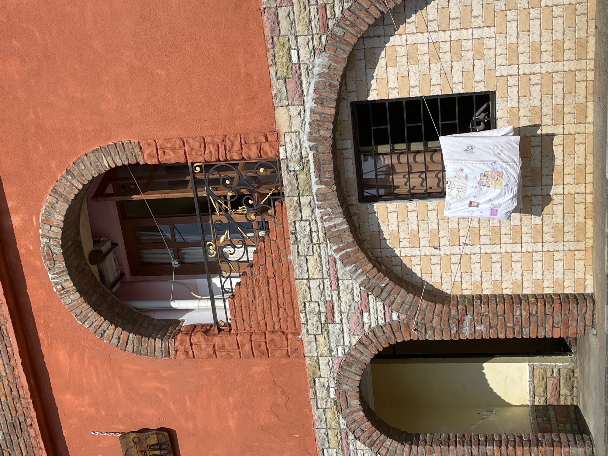 This media file shows an exterior view of a building's facade, focusing on architectural details and features. The building displays a mix of materials, including a rust-colored stucco on the left and a patterned tile facade on the right, composed of cream, yellow, and pink rectangular tiles. Both sections are framed by arched brickwork.
On the left side, a curved opening reveals a window with dark wooden frames and a decorative black metal grate. Inside the opening, there are hints of interior space, including a white pipe. A small, carved wooden object with a metal chain hangs on the stuccoed wall below this opening.
On the right side, a rectangular window is visible behind a black metal grate. A white duvet or blanket, decorated with cartoonish animals in pink and yellow, is hung on a thin wire across the window, suggesting laundry is being dried. The patterned tile wall extends further down, framing another arched opening at the bottom, which shows a glimpse of a yellow-painted interior space.
The lighting suggests it is daytime, with strong sunlight casting shadows, particularly on the lower arched opening. The scene appears to be in a location with a warm climate, possibly Ureki, Georgia, given the context. There are no people visible in the image. The overall impression is one of a residential building with unique architectural design elements.