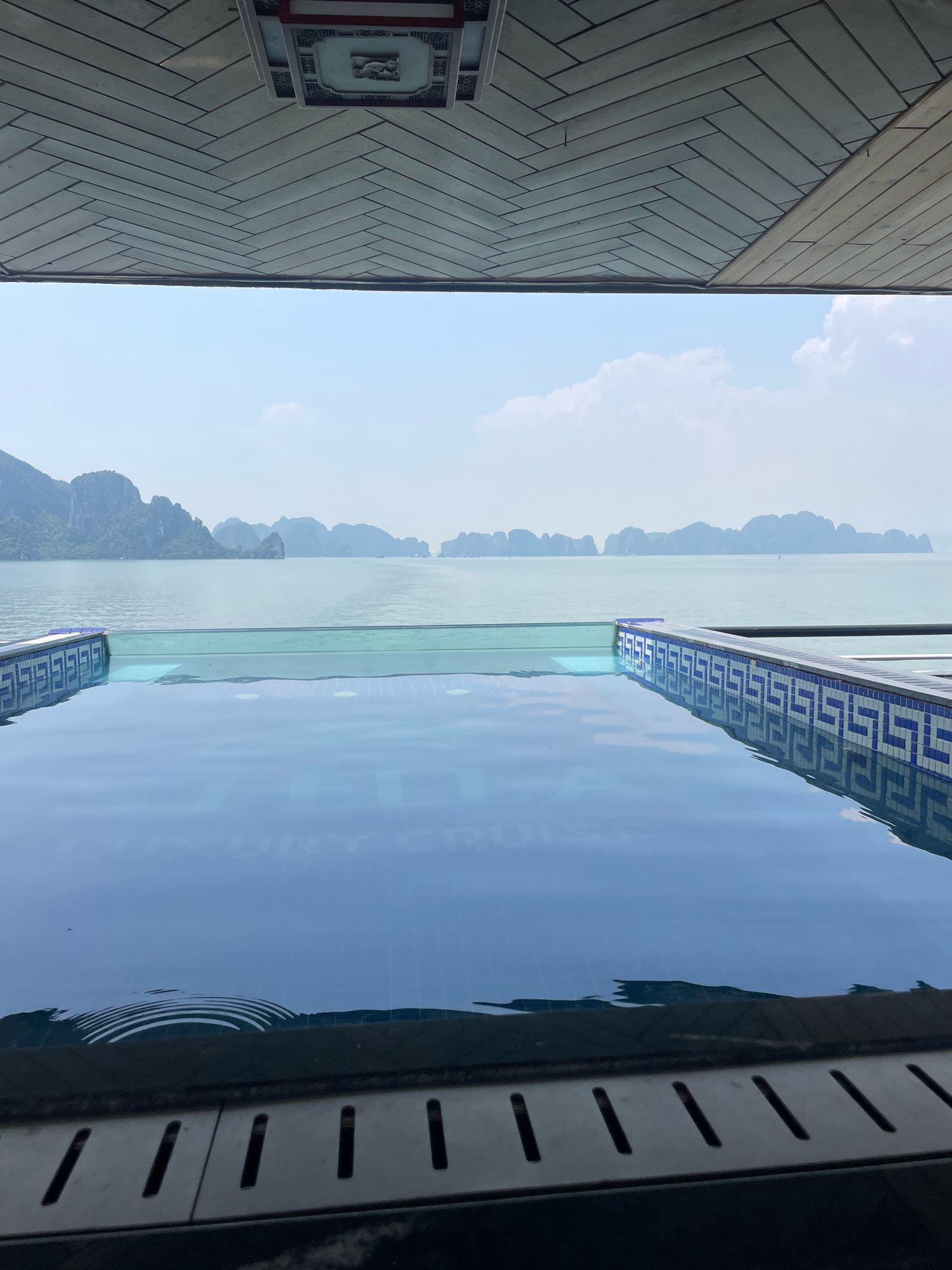 The image depicts an infinity pool overlooking a body of water with numerous limestone karsts and islands, characteristic of Halong Bay, Vietnam. The pool features a blue and white tiled edge and its surface reflects the sky and the distant landscape. In the foreground, a metal grate is visible. The sky is partly cloudy, and the water appears calm. Above the pool, a portion of the ceiling is visible, adorned with a patterned design and a decorative element. The overall scene conveys a sense of relaxation and scenic beauty.