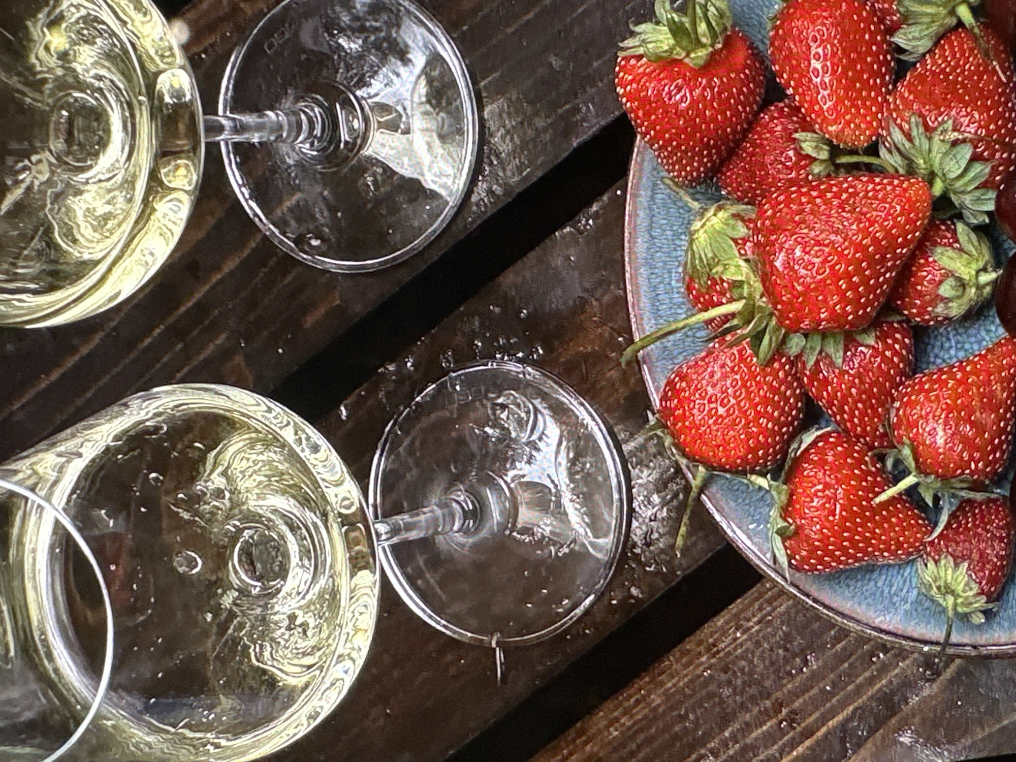 A still life photograph depicts two wine glasses filled with a light-colored liquid, presumably wine, positioned beside a blue plate heaped with ripe strawberries. The setting appears to be a rustic, dark-stained wooden surface, potentially an outdoor table or deck, with visible water droplets suggesting recent rain or condensation. The composition is arranged at an angle, offering a slightly asymmetrical view. The likely location is not discernible from the provided visual information; there is no immediate indicator of Yerevan, Armenia, unless supplementary contextual cues are available.