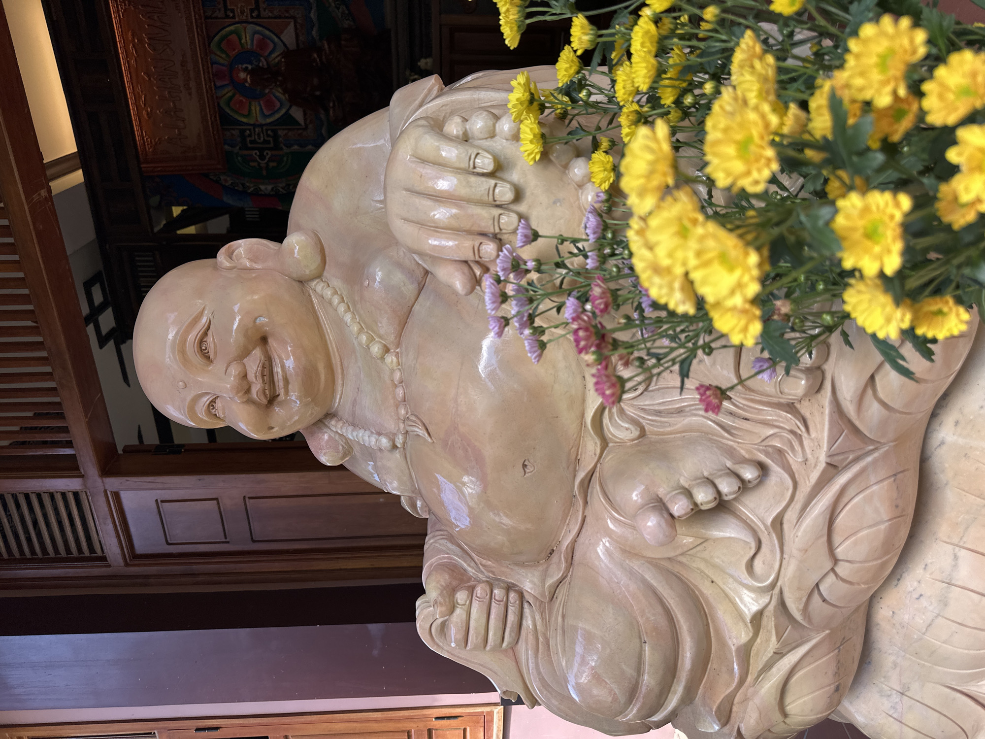 The image shows a statue of a smiling, bald man, likely a depiction of Budai or the Laughing Buddha, a figure associated with good luck and prosperity in Chinese Buddhism. The statue is made of a light-colored, smooth material, possibly stone or ceramic, with a glossy finish. It is adorned with a beaded necklace. The statue is seated, with its hands resting on its lap and its legs crossed. The figure has a jovial expression, with rosy cheeks and a wide smile.
A vibrant bouquet of yellow and pink chrysanthemums is placed in front of and partially obscuring the statue, adding a touch of color and life to the scene. The flowers are in full bloom, with their petals fanned out. The background suggests an indoor setting, possibly a temple or a richly decorated room, with wooden paneling and ornate decorative elements visible. There is a framed piece of artwork with colorful patterns and some text in a language that appears to be Vietnamese on the wall behind the statue. The overall impression is one of tranquility and warmth, evoked by the cheerful demeanor of the statue and the presence of fresh flowers. The lighting suggests it could be daytime, with the ambient light highlighting the statue's features and the flowers. The location context provided, Thôn 5, Vietnam, indicates a Southeast Asian cultural setting.