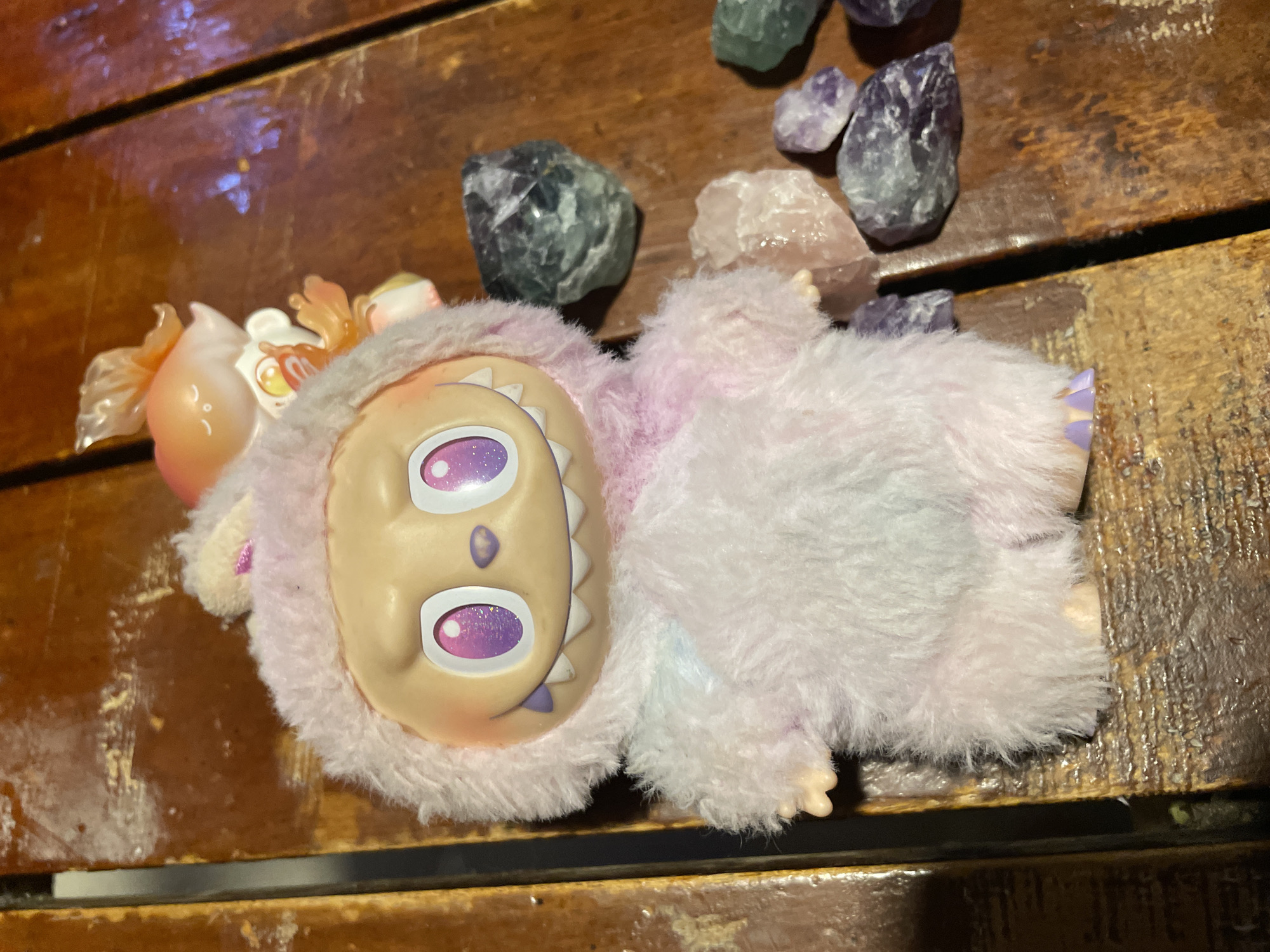 A pink and white fluffy plush toy with a light tan face, large glittery purple eyes, and a wide smile displaying white teeth and a purple gumline stands centered on a dark brown wooden surface. The toy has small, pointed ears on its hood and small, light-colored hands and feet, with the feet showing purple tips. Behind the plush, slightly to its upper right, a smaller, translucent orange figurine with elaborate, fin-like details is partially visible. To the right and front of the plush, several unpolished, irregularly shaped mineral or crystal specimens are scattered across the wooden surface. These specimens exhibit colors including dark green, light pink, and various shades of purple. The wooden surface, composed of multiple planks, displays a glossy finish and visible grain lines. No human figures are present. All objects are stationary. The scene is located in Davao City, Philippines.