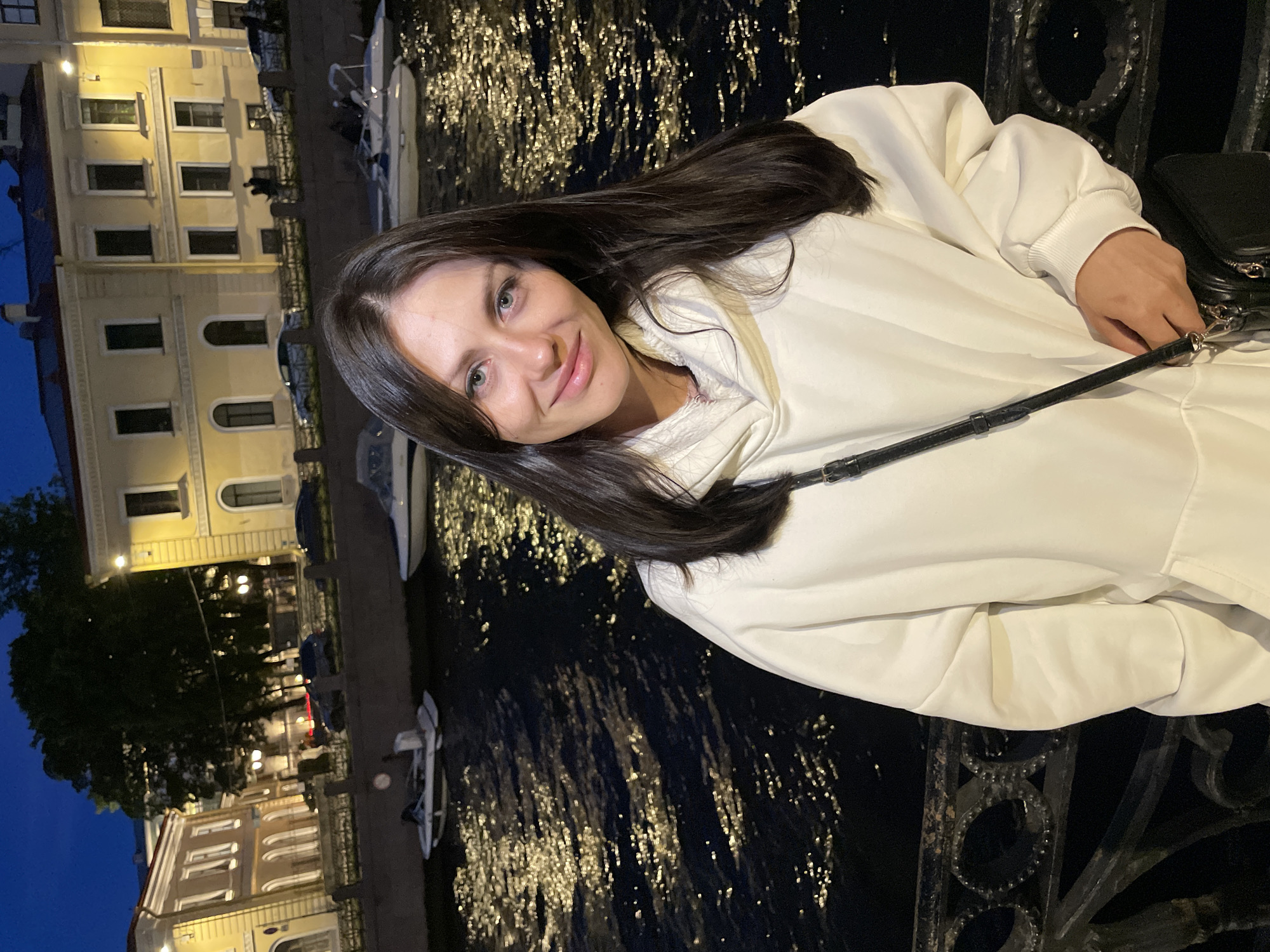 A young woman with long, dark hair and blue-green eyes is standing on a street in Saint Petersburg, Russia, at dusk. She is wearing a loose-fitting white hooded sweatshirt and a black crossbody bag. The woman is smiling at the camera, with her left arm bent and resting on a dark, ornate metal railing. Behind her, a dark canal reflects the lights of the city, and several boats are docked along the bank. Across the canal, there are two-story buildings with yellow facades and arched windows, illuminated by streetlights. The sky is a deep blue, indicating the transition from day to night.