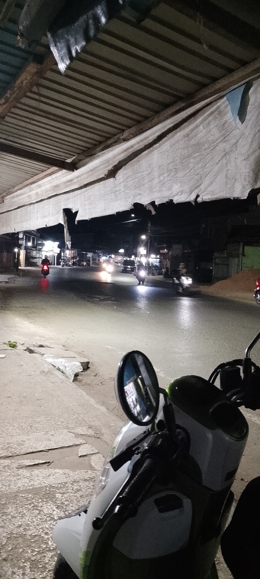 The image captures a nighttime street scene in Tungkaran Pangeran, Indonesia, seemingly under an overpass or a structure with a corrugated metal roof and hanging white fabric. The foreground is dominated by the front section of a white and green scooter, with its handlebar and side mirror clearly visible. The mirror reflects the street scene. The road ahead is illuminated by the headlights of vehicles, including motorcycles and cars. Several motorcycles are in motion, one with a red taillight in the distance to the left, and another with bright headlights in the middle ground to the right. Buildings line the street on both sides, some appearing to be shops or residences with lit windows. The pavement is wet, reflecting the artificial lights. A concrete sidewalk with some debris, including plastic bags, is visible to the left of the road. The overall atmosphere is that of a typical busy street at night in an urban or semi-urban environment.