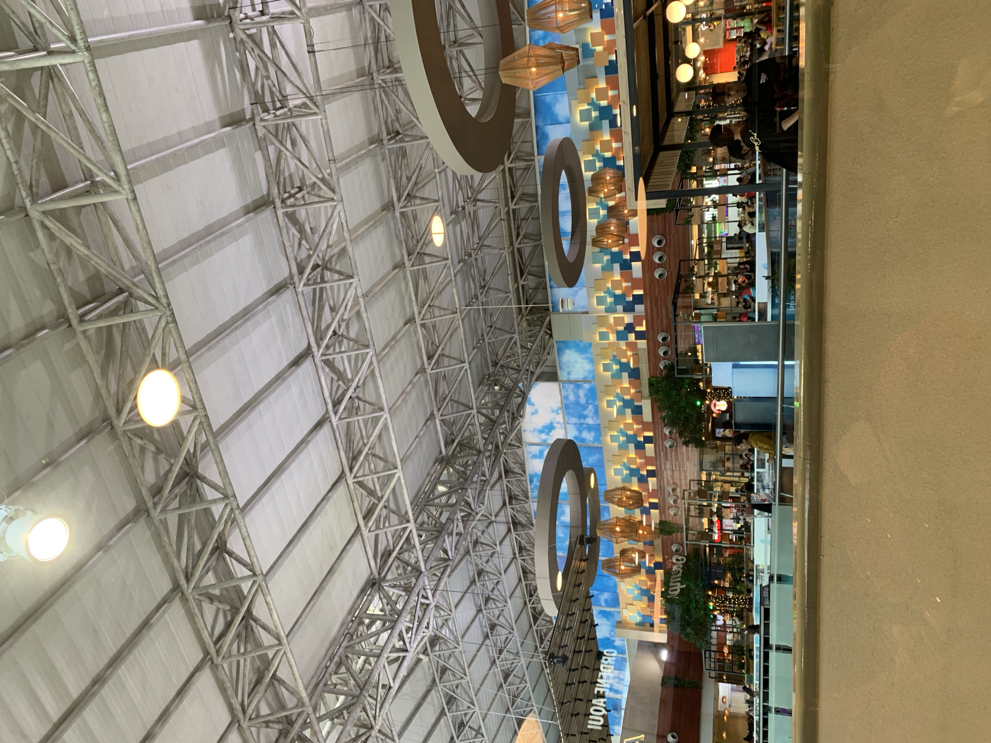 The image shows the interior of the Sambil Margarita shopping mall in Porlamar, Venezuela. A large metal structural grid forms the roof, with evenly spaced lights. Circular decorative rings with pendant lamps hang below the roof. A wall with a blue and brown tiled design is visible on the right side. People are visible on the second level of shops and restaurants. A wall with a railing is visible in the foreground, partially obscuring the scene.