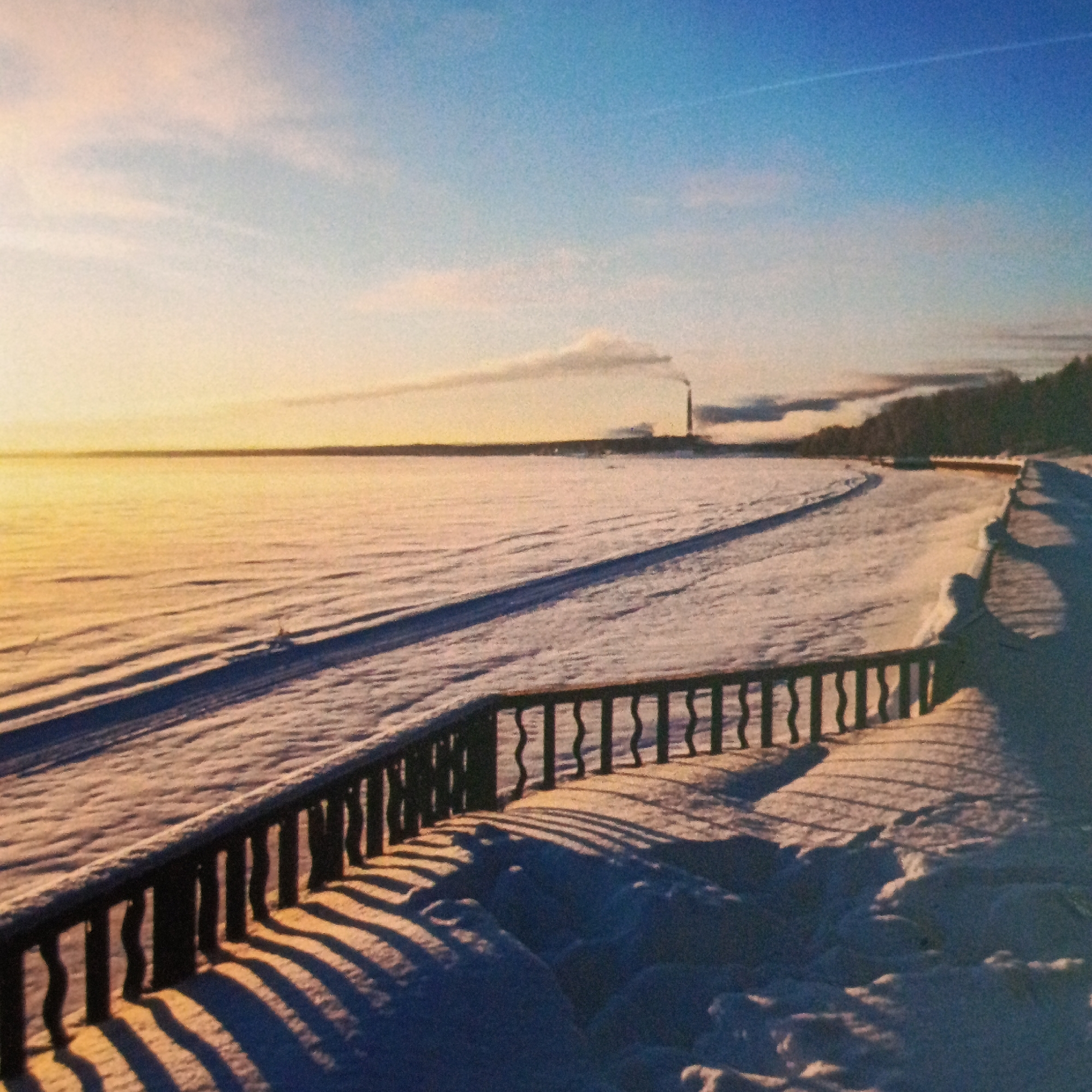 The image depicts a snow-covered landscape in Марковский, Russia, under a sky transitioning from blue to yellow-orange, suggesting dawn or dusk. A rail fence runs along the foreground, casting shadows on the snow-covered ground. Beyond the fence, a vast expanse of snow stretches to the horizon, possibly a frozen lake or field. In the distance, there is a structure resembling a smokestack emitting smoke, and a treeline is visible on the right side of the frame.