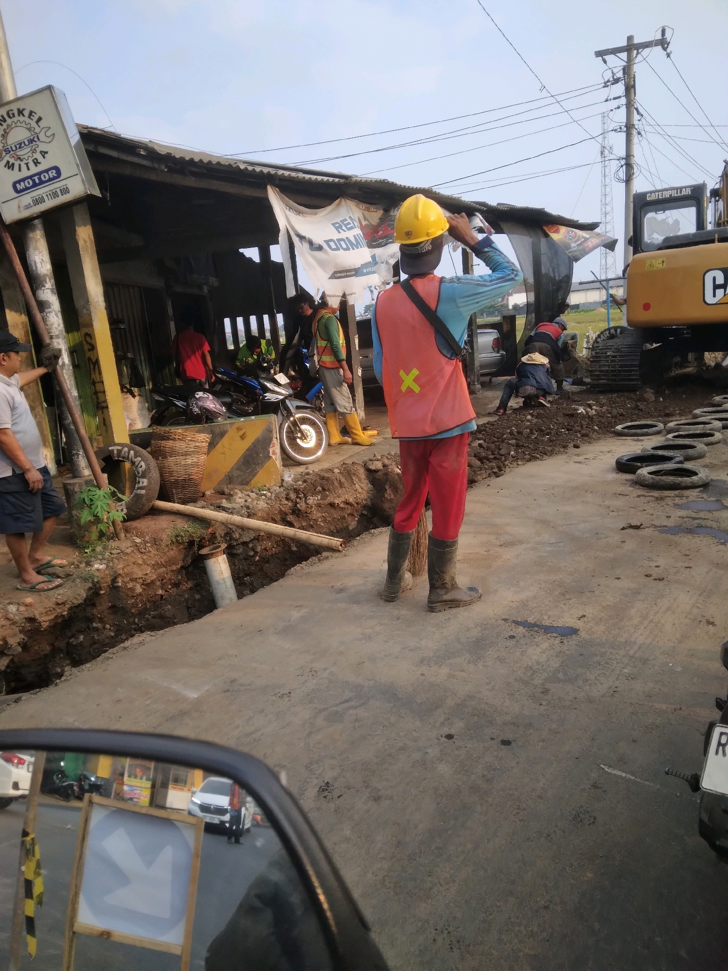 In Desa Pamijen, Kecamatan Sokaraja, Indonesia, a construction scene unfolds with workers and machinery on a sunlit day. A man wearing a yellow hard hat, orange safety vest over a blue long-sleeved shirt, and red pants stands facing away from the camera, with his right hand near his head. He wears dark boots. In the background, several other construction workers are visible. One individual, also in an orange safety vest and blue shirt, is bent over near a yellow excavator. Another person, wearing a straw hat and blue clothing, is similarly occupied. A large yellow excavator with "CATERPILLAR" branding is positioned to the right. Several black tires are arranged in a line on the ground. The scene includes a deep trench dug into the earth, with exposed soil and debris. To the left, a building with a corrugated metal roof is visible, along with parked motorcycles. A sign for "SUZUKI MITRA MOTOR" is mounted on a pole. Another man in a light-colored shirt and dark shorts stands near a pole on the far left, looking towards the construction area. A motorcycle mirror in the foreground reflects a white car and a directional sign. The ground is a mix of concrete and dirt, with some puddles of water. Overhead, power lines are strung between utility poles.