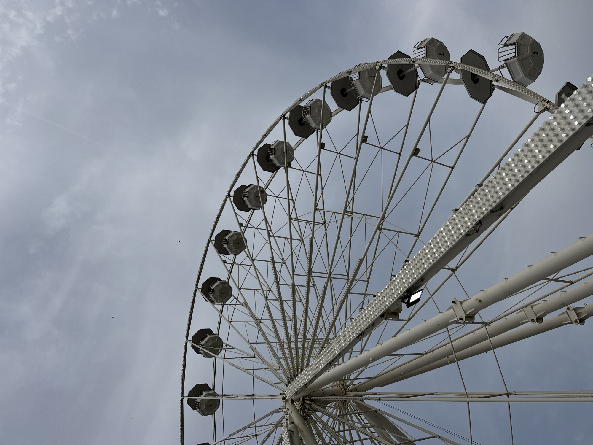 The image depicts a large Ferris wheel, captured from a low angle against a cloudy, overcast sky. The Ferris wheel is white, with enclosed passenger cars attached to its outer rim. These cars are grey with a partly open design. White lights are mounted along the supporting structure of the wheel. The Ferris wheel is located in Plovdiv, Bulgaria. The composition emphasizes the scale and structure of the Ferris wheel against the sky. There are a few birds flying in the background.