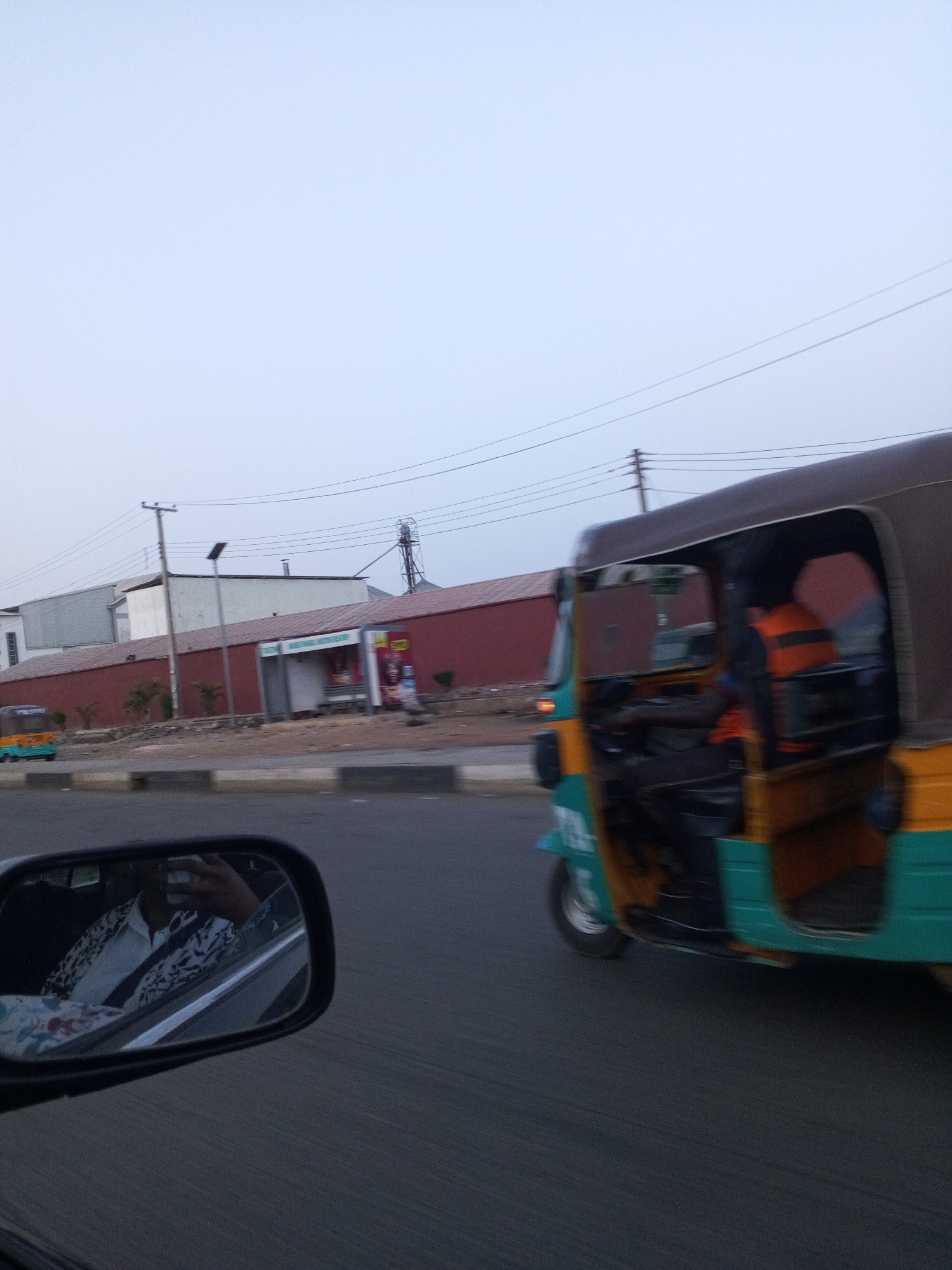 The image captures a roadside scene in Jos, Nigeria, from the perspective of a moving vehicle, evident by the reflection of a person in a side mirror. The sky is overcast and bright, suggesting either early morning or late afternoon.
In the mid-ground, a "keke napep" (motorized tricycle) is prominently featured, moving alongside the photographer's vehicle. It has distinctive yellow, green, and brown panels. Inside, the driver is visible, wearing an orange and black striped garment, possibly a safety vest or shirt. The road beneath both vehicles shows motion blur.
To the left of the road, a concrete barrier separates lanes, beyond which lies a dusty, unpaved strip with sparse vegetation. Further back, a long, reddish-brown wall runs horizontally across the frame. Behind this wall, several industrial-style buildings, predominantly white and grey, are visible. A tall metal structure, possibly a silo or part of a factory, rises in the background. Utility poles with power lines crisscross the upper left portion of the sky, and a solar-powered street lamp stands near the wall. Another yellow and green tricycle is visible in the distant left.
Along the red wall, an advertising shelter resembling a bus stop is present. Text on the shelter clearly reads "PREMIER FLOUR MILLS" in white letters on a green background, with other indistinguishable words and product images. The person reflected in the side mirror is wearing a patterned long-sleeved shirt over a white collar, and appears to be holding a phone, likely taking the photograph.