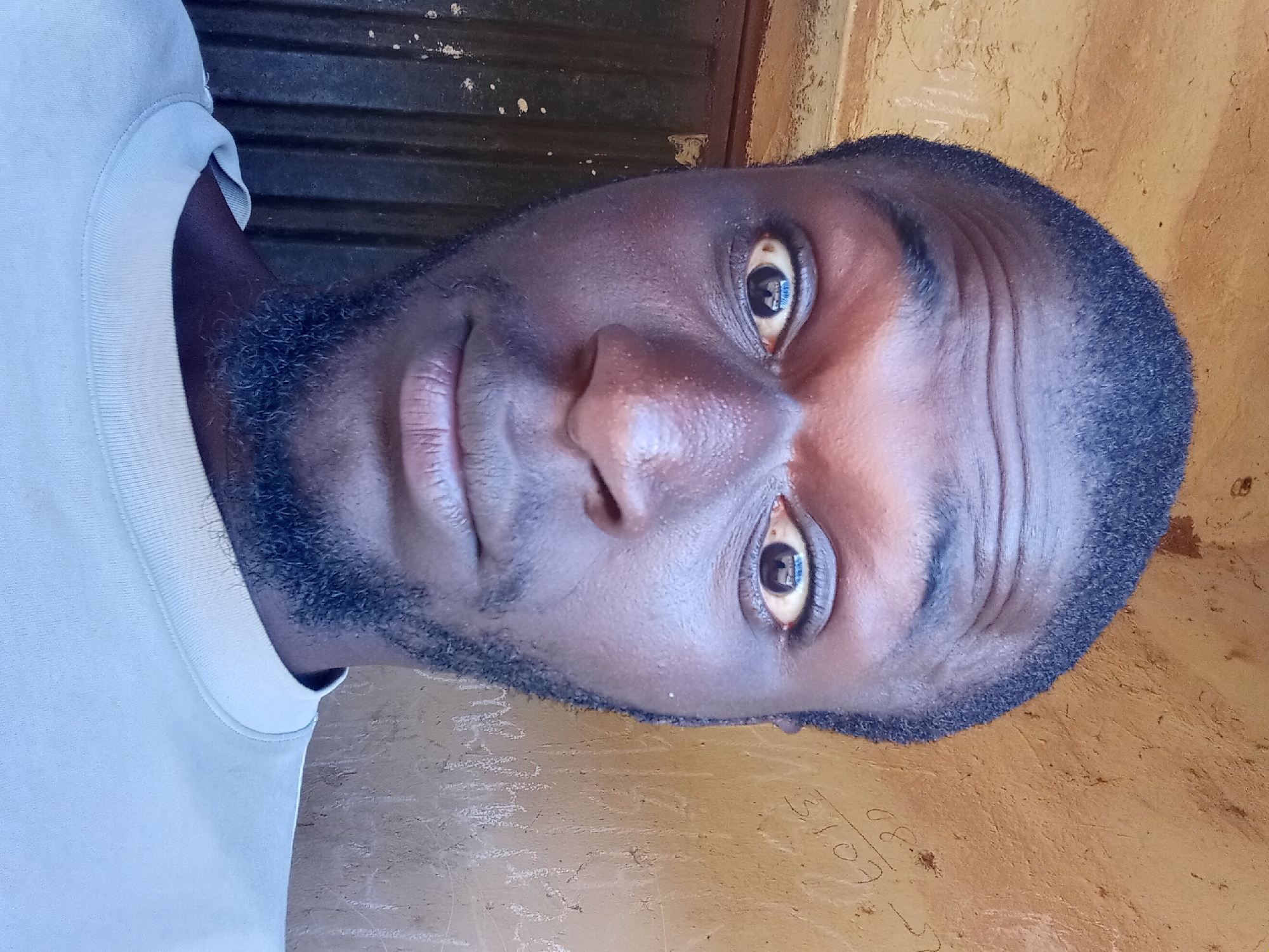 A selfie shows a close-up of a Black man's face and upper torso, set against a textured, ochre-colored wall with some faint white markings. The man has short, dark hair and a short, dark beard. His eyes are wide open, and his eyebrows are raised, giving him an alert or surprised expression. He is wearing a light-colored t-shirt with a ribbed neckline. The background wall appears to be outdoors, possibly part of a building in Bukuru, Nigeria, given the context. To the left of the man's face, a dark, corrugated metal surface is visible, possibly a door or a wall panel. The lighting suggests it is daytime.