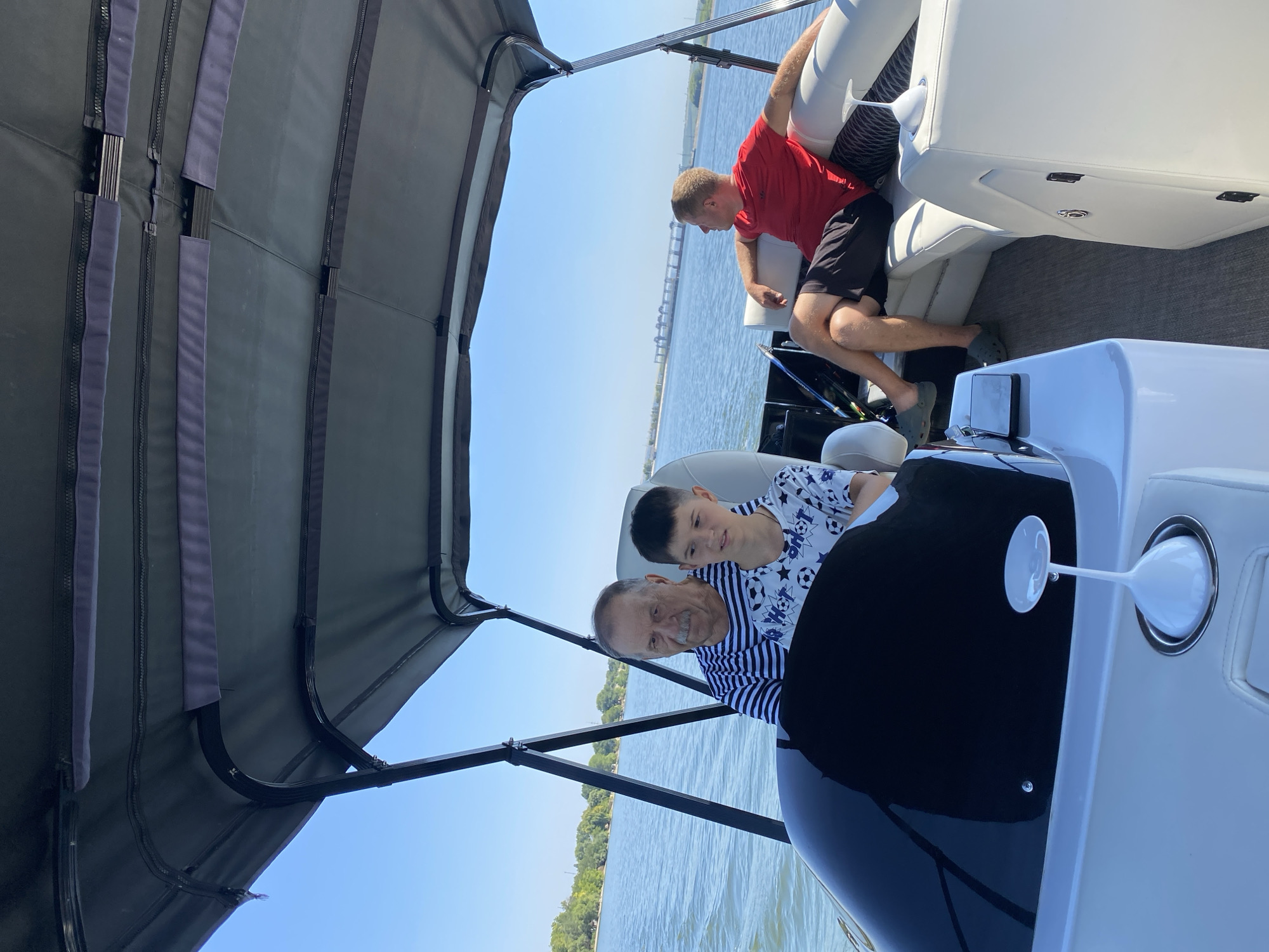 The image shows three individuals on a boat on a body of water. In the foreground, a man with gray hair and a mustache, wearing a striped shirt, is at the helm. Next to him, a young boy in a t-shirt with a soccer ball pattern looks forward. In the background, a man wearing a red shirt and black shorts is seated facing away from the camera, looking towards the horizon. The boat has a dark canopy overhead and white seating. The water is blue, and the sky is clear. Trees are visible on the distant shore. A bridge-like structure is in the far distance. The scene appears to be taking place on the Dniester River near Korzhevo, Moldova, given the specific location requested.