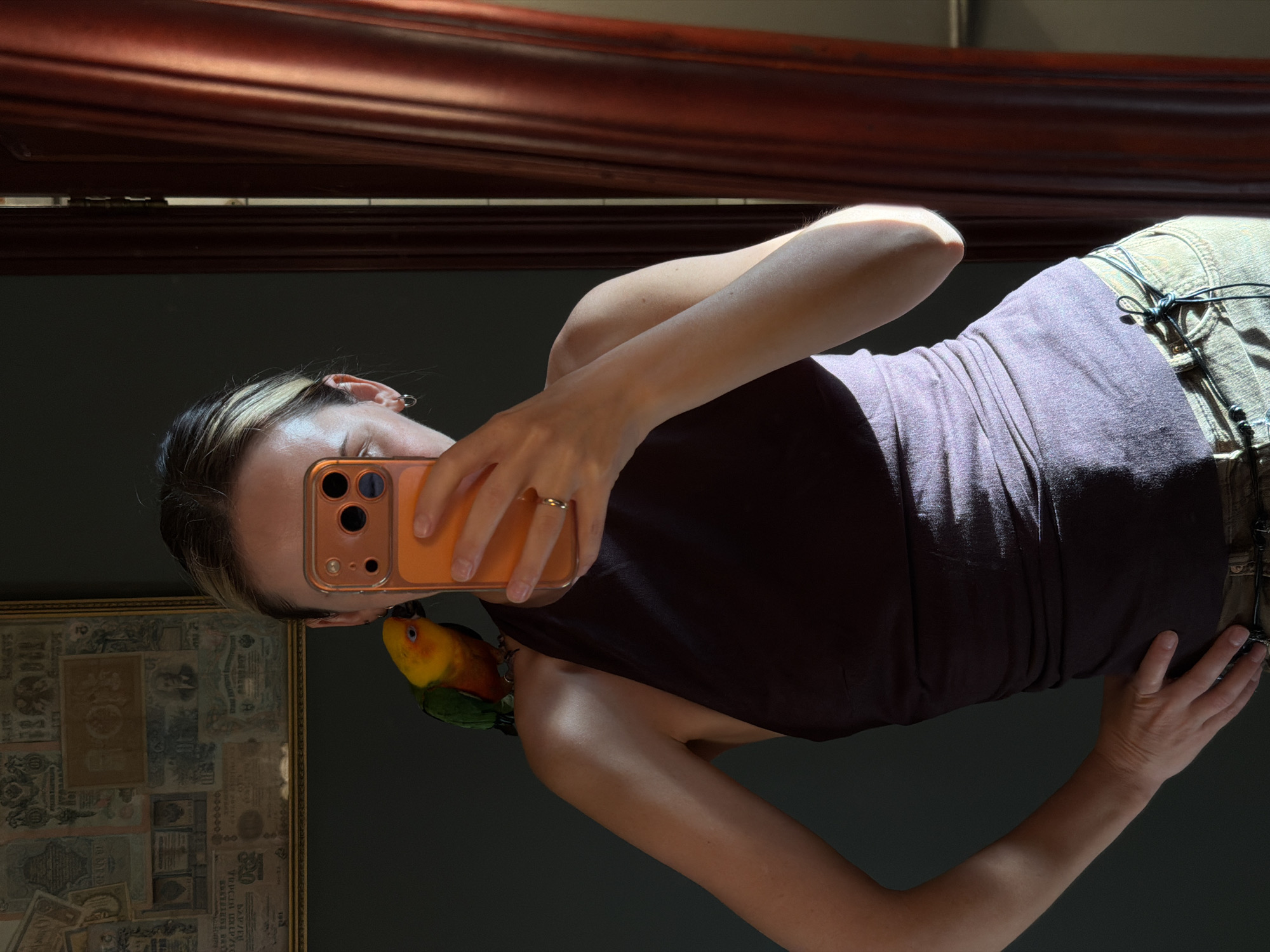A woman takes a selfie in what appears to be a dimly lit room, possibly a bedroom, with sunlight streaming in from the right. She is wearing a dark purple tank top and light-colored pants. A small, colorful parrot is perched on her left shoulder, near her face. The woman is holding an orange smartphone in her right hand, angled to capture the reflection in a mirror. The background includes a wall with framed artwork, a dark green wall, and a portion of a red curtain. The woman's hair is pulled back, and she is wearing a hoop earring in her visible ear and a ring on her right hand. The overall lighting suggests it is daytime, with strong directional light creating highlights and shadows. The image does not contain any information that would allow for identification of the location as Khimki, Russia.