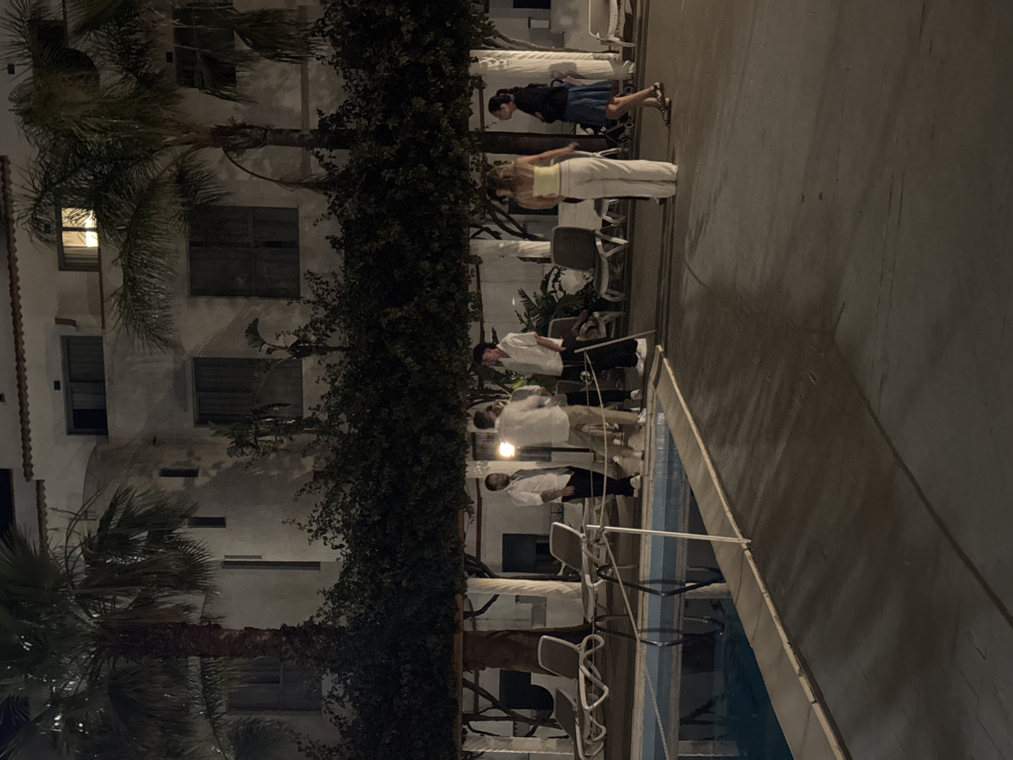 The image, rotated vertically, depicts an outdoor scene at night, likely within a resort or hotel complex in Isola delle Femmine, Italy. Artificial lighting provides illumination for the area, which includes a swimming pool, a multi-story building, and various plants.
In the mid-ground, two women are visible. One wears a light-colored top and white pants, while the other is dressed in a dark top, a dark blue skirt, and patterned shoes. Closer to the blue swimming pool on the right, a group of three to four men, possibly staff, are present, uniformly dressed in white shirts and dark trousers. One man appears to be positioned behind a counter or bar area, suggesting service activity, while others stand near the pool's edge.
The background on the left features a white-walled building with multiple windows, some fitted with dark shutters. A single window on a lower floor emits a warm light from within. Tall palm trees and dense, dark green foliage partially obscure the building. The foreground around the pool area contains several outdoor chairs and tables, some of which appear stacked or arranged. The swimming pool's blue water reflects the dim ambient light, and a white rope or railing is visible along its edge. The scene conveys a calm, evening atmosphere typical of a Mediterranean resort. No visible text is present.