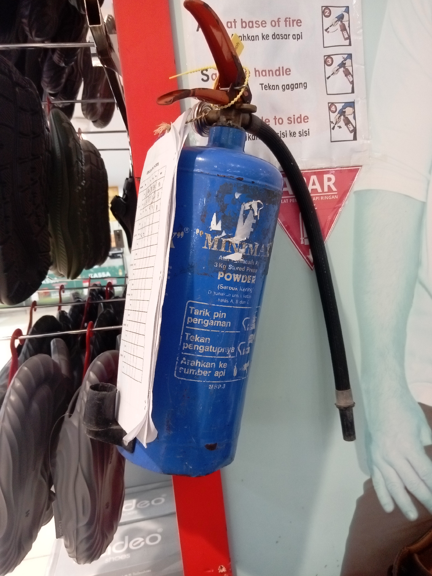 A blue fire extinguisher hangs from a metal rack with red support beams. Attached to the extinguisher is a white inspection tag with a grid of handwritten entries and dates. The fire extinguisher has a black hose attached to its nozzle. Above the extinguisher, a sign with instructions for its use is partially visible. The sign has text in both English and Indonesian, including phrases like "at base of fire" and "handle". To the left of the fire extinguisher, several pairs of black shoes are hanging from hooks. In the background, a poster with the word "AR" is visible, along with a faint outline of a person's arm and hand. The overall scene suggests a commercial or storage environment, possibly a retail store in Batam, Indonesia, given the Indonesian text on the sign.