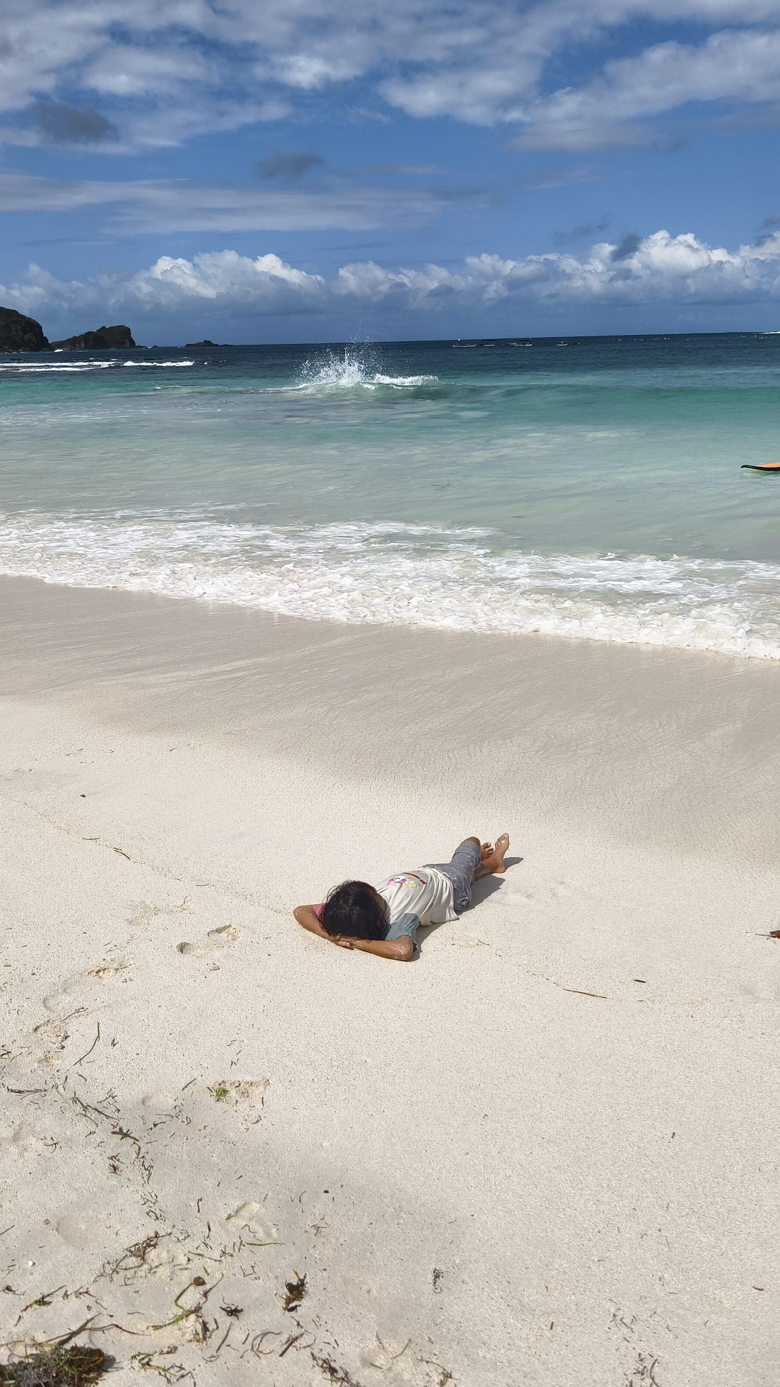 A bright, vertical outdoor photograph captures a serene beach scene, likely in Indonesia, under a clear blue sky with scattered white cumulus clouds. In the foreground, a child or small person is lying face down on the expansive, light-colored sand beach, resting their head on their folded arms. They are wearing a light-colored t-shirt with a faint design on the back and dark pants or shorts, with their bare feet visible. Footprints are subtly visible in the sand near them, and some dark seaweed or natural debris is scattered in the lower left corner.
The middle ground features the clear turquoise and blue ocean, with gentle white-capped waves breaking near the shore. One wave further out creates a noticeable splash. A small, partially visible light brown or orange object, possibly a surfboard, can be seen at the far right edge of the water. In the distance, dark, rocky hills or formations extend from the coastline on the left, and several small boats are faintly visible on the horizon line. The scene conveys a sense of peaceful relaxation on a sunny day. No visible text is present.