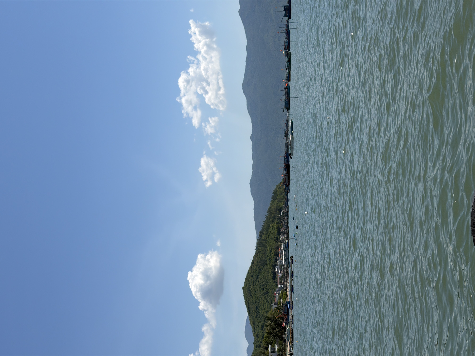 This is a wide-angle, eye-level shot of a coastal landscape in Ninh Ích, Vietnam. The foreground is dominated by a body of water with moderate ripples, reflecting the bright, overcast sky. In the mid-ground, several small boats and buoys are scattered across the water, some appearing to be part of a fish farm or aquaculture operation. A coastline with green, tree-covered hills runs across the horizon. Along the base of the hills, a settlement of buildings and a pier are visible. A line of larger boats, possibly fishing vessels, is anchored in the distance. The sky above is a clear blue with scattered white clouds.