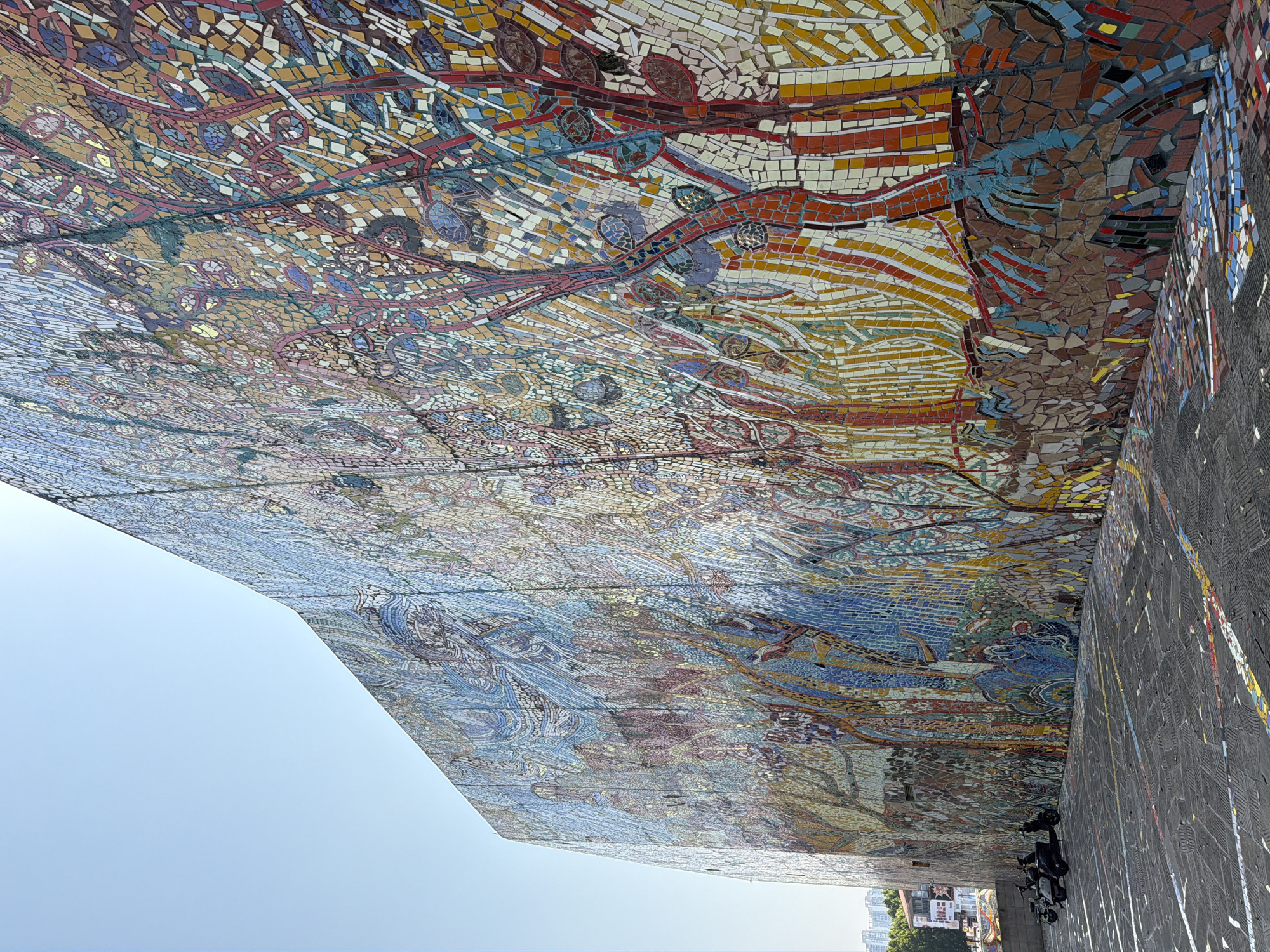 This image depicts a vast, vibrant mosaic mural on the side of a building in the Chongqing High-tech Zone, China. The mural covers the entire visible surface of the building, extending from the foreground up to the sky. The artwork is composed of numerous small, irregularly shaped tiles in a wide array of colors, including blues, greens, yellows, oranges, reds, and browns. The design appears to be abstract or stylized, featuring organic shapes, possibly representing trees, foliage, or flowing water. The mosaic is highly textured and catches the sunlight, creating a shimmering effect.
In the foreground, a paved area is visible, which appears to be a public space or walkway. The paving stones are dark and have colorful tile fragments embedded within them, mirroring the mural's palette. Three scooters are parked on the paved area, to the left of the frame. They are dark in color and appear to be stationary.
The sky above is clear and a pale blue, indicating daylight. In the far background, on the left side of the image, hints of other buildings and greenery can be seen, suggesting an urban or semi-urban environment. The perspective of the photograph emphasizes the immense scale of the mosaic artwork.