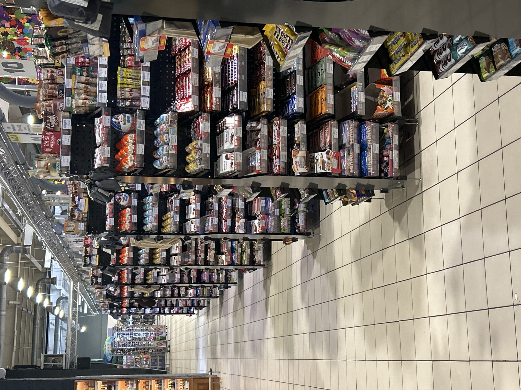 The image depicts an aisle in a supermarket, specifically showing a display of confectionery and beverages. The aisle features multiple shelves stocked with various brands of chocolate bars, candy eggs, coffee products, and other packaged snacks. Notable brands visible include Kinder, Snickers, Nescafe, and various other items that appear to be chocolates and sweets. The shelves are organized in a typical retail manner, with products displayed in their original packaging. The flooring is tiled with a light-colored ceramic tile. Overhead lighting fixtures are visible, illuminating the aisle. In the background, other aisles with more products and shoppers can be seen, suggesting a busy retail environment. The overall impression is that of a well-stocked supermarket confectionery section. The image is dated as being taken in Brovary, Ukraine.