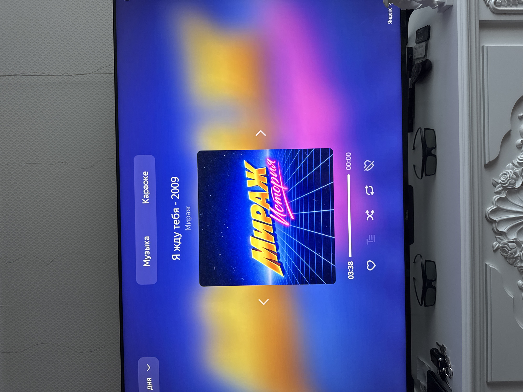 The image displays a television screen showing a music interface. The screen features a song title "Я жду тебя - 2009" (I'm waiting for you - 2009) by an artist or group named "Мираж" (Mirazh). The album art visible is in a retro, neon-grid style with the text "МИРАЖ История" (Mirazh History). A progress bar indicates the song is at 03:38 out of 00:00, suggesting it is near the end. Playback controls, including a heart icon for liking, and other icons for repeat, shuffle, and a cross symbol, are present at the bottom of the screen. The background of the screen is a gradient of blue, yellow, and pink. On the shelf beneath the television, a pair of 3D glasses, a set of keys, and several remote controls are visible. The wall behind the television has a textured wallpaper pattern. There is no indication of the city of Oskemen, Kazakhstan, in the image.