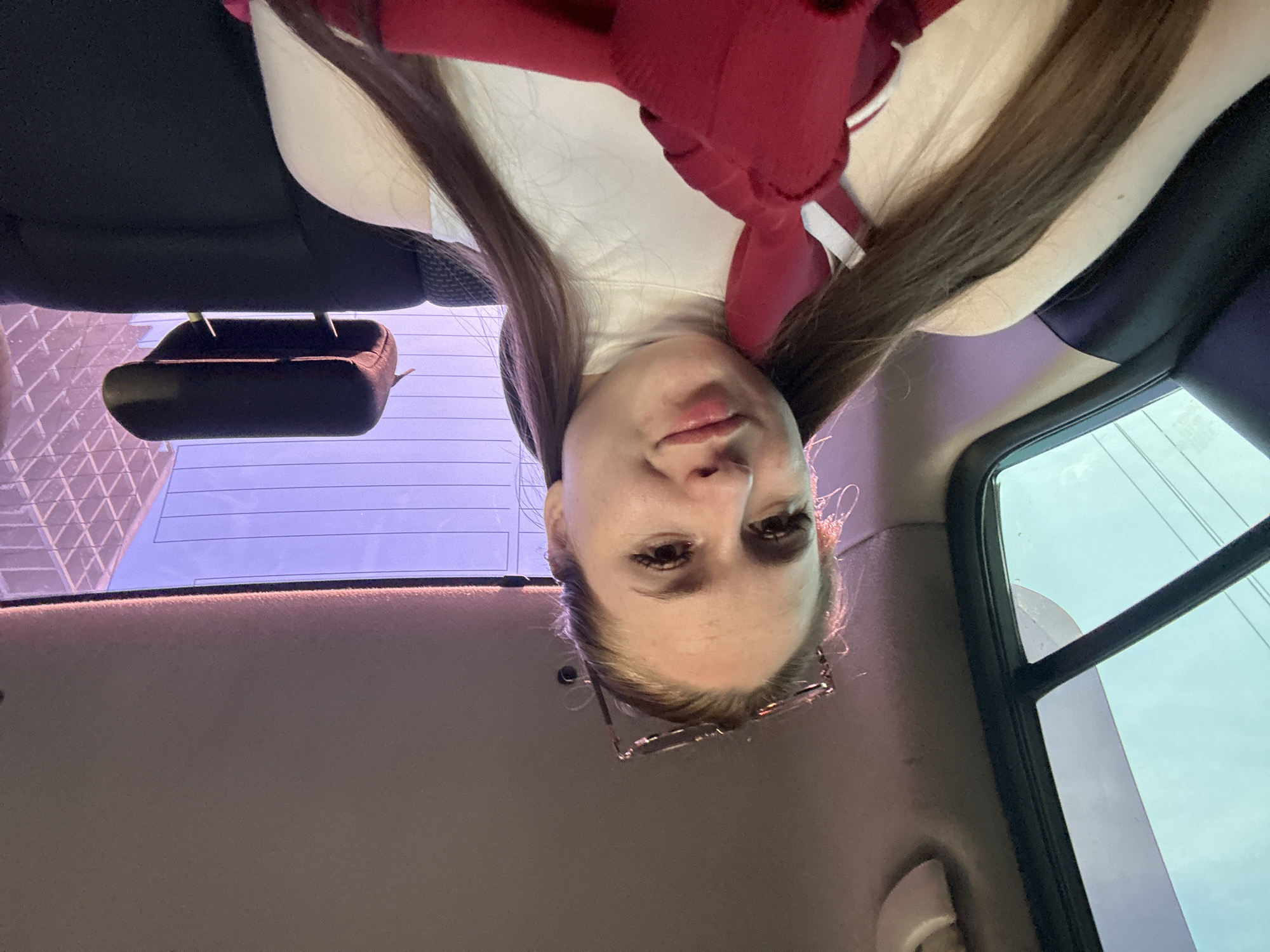 A young adult female with fair skin and long, straight brown hair pulled back, wearing a white high-neck sleeveless top, is seated in the rear passenger area of a vehicle. She holds a red garment with white stripe detailing around her neck and chest. Glasses are perched on her head. She looks directly at the camera with a subtle smile. The vehicle's interior features a light-colored headliner, black seat upholstery, and a thin red ambient light strip along the roofline. Through the left rear window, a light blue sky with faint power lines is visible. The rear windshield displays a purplish sky and a modern building characterized by a distinct geometric, grid-like facade, along with horizontal defroster lines. The scene is located within a city, in the city of Unknown, Unknown.