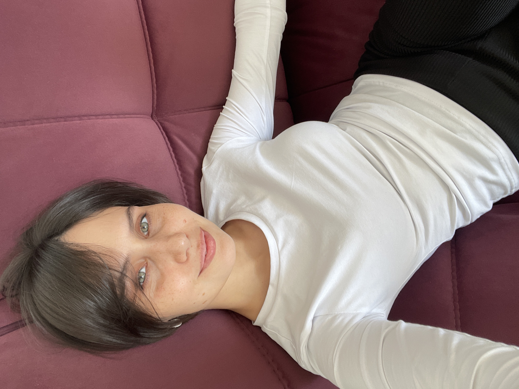 A woman with dark hair and green eyes is lying on a purple couch. She is wearing a white long-sleeved shirt and black pants. The location is Izhevsk, Russia.