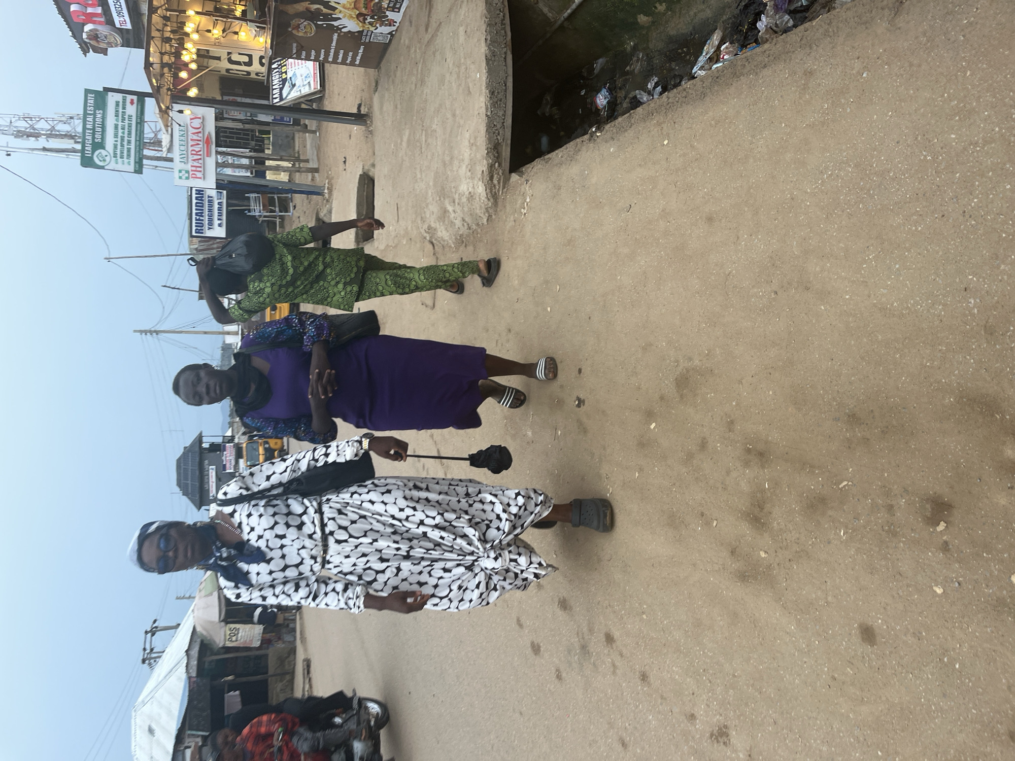 The image depicts a street scene in Abuja, Nigeria, characterized by a dusty, unpaved road and a row of small shops. The sky is overcast, suggesting a cloudy day. In the foreground, three individuals are walking on the dirt road. The person in the center is a woman wearing a long, black and white polka dot dress, sunglasses, and a headscarf. She is carrying a black bag on her shoulder and holding a small, black umbrella. To her right, another woman walks in a purple dress, and to the left, a person in a green patterned outfit is walking away from the camera, carrying a black plastic bag over their shoulder. Further in the background, there are more buildings, a motor tricycle (Keke napep), and various signs for businesses, including "LEAFGATE REAL ESTATE SOLUTIONS" and "JAYCEEKEL PHARMACY". The overall impression is that of a busy, everyday street in an urban area of Nigeria, with some visible signs of commercial activity and the presence of informal settlements. The presence of a drainage ditch filled with trash on the right side of the image suggests some issues with waste management.