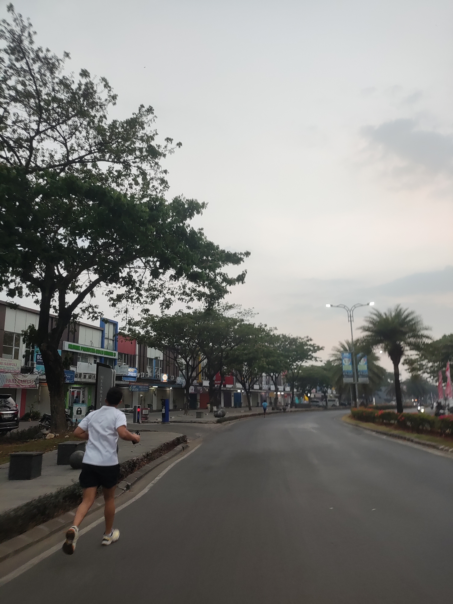 A man runs on the right side of a road in a city. He is wearing a white t-shirt and black shorts. He is running away from the camera, and his body is angled to the left. The road is paved and there are trees on the left side of the road. There are also buildings on the left side of the road. The sky is overcast. The image appears to be taken at dusk. The man is running on a paved road that has a curb and a sidewalk. The sidewalk has some planters and benches. The buildings on the left are commercial establishments. The man is running on the right side of the road, which is typically the direction of traffic in Indonesia. The image depicts an urban environment in Telagaasih, Indonesia, with a man engaged in outdoor exercise.
