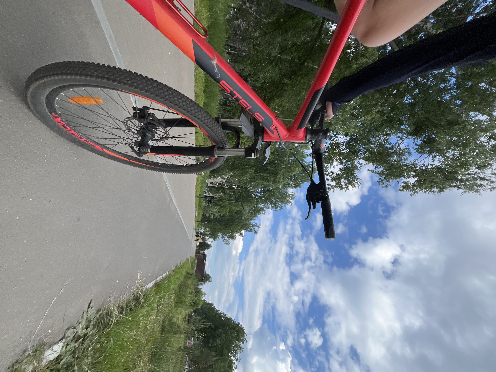 The image shows a first-person perspective of a person riding a red and black Stels bicycle on a paved bike path in Chaikovsky, Russia. The bicycle's front wheel and a portion of the frame are prominently in the foreground. The rider's left arm, wearing a dark long-sleeved shirt, is visible on the handlebar. The bike path stretches into the distance, lined with green trees and grass. The sky is blue with scattered white clouds. In the background, several other people are seen walking along the path, and a small building is visible amongst the trees. The overall environment appears to be a park or recreational area within the city.