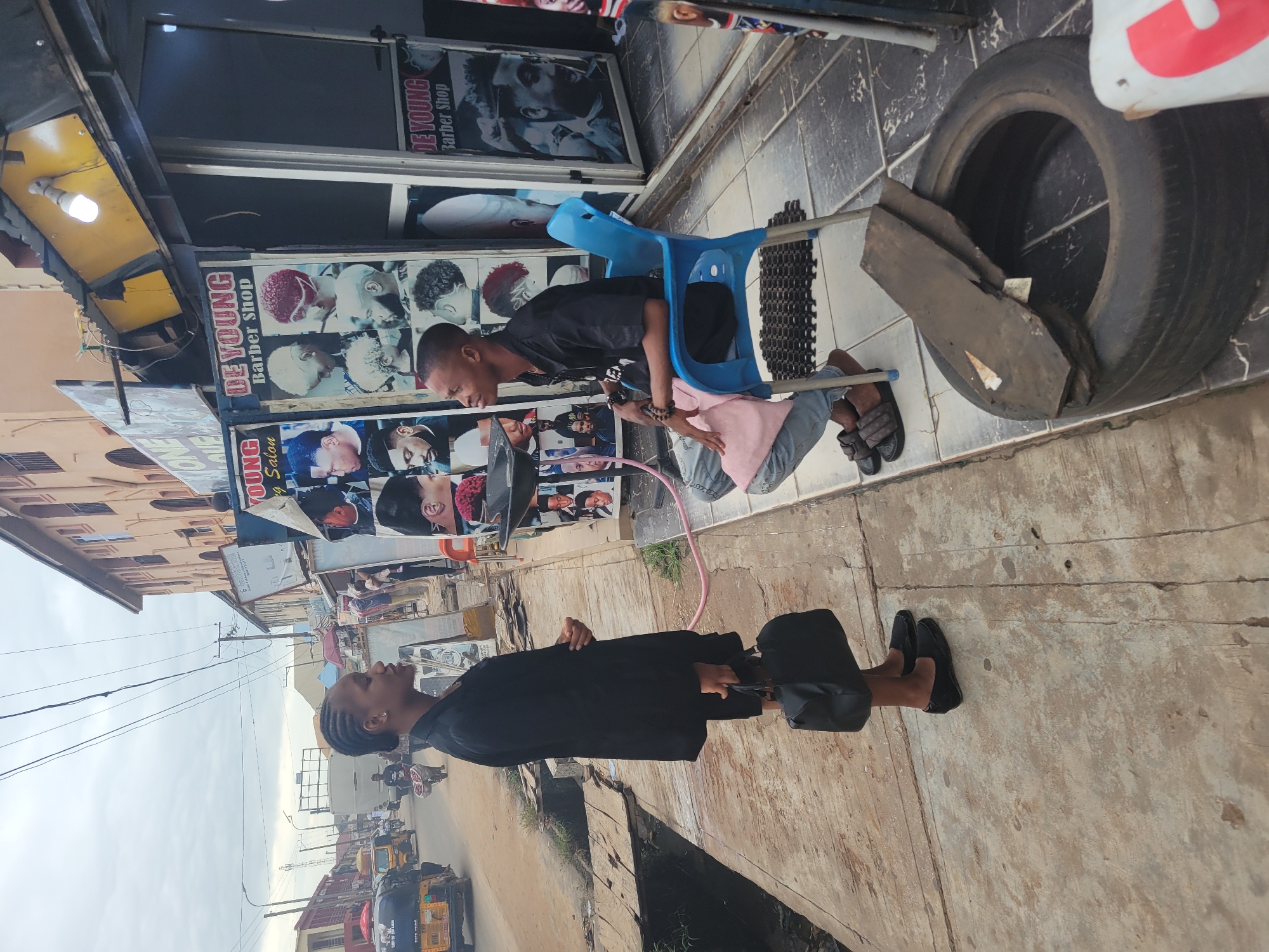 The image depicts a street scene in Egbagu, Nigeria. Two young people are the primary subjects. In the foreground, a person wearing a black outfit and black loafers stands facing away from the camera, holding a black bag. Behind them, another person sits on a blue plastic chair in front of a barber shop. This individual is wearing a black t-shirt, light blue jeans, and dark sandals, and is looking towards the person standing.
The barber shop features posters of hairstyles on its facade. Visible text includes "DE YOUNG Barber Shop". The street appears to be paved with concrete slabs, and there is a discarded tire with some debris inside. The setting suggests an urban or semi-urban environment, with buildings and a street visible in the background, including some three-wheeled vehicles that appear to be auto-rickshaws. The sky is overcast, indicating a potentially cloudy day. The overall mood appears casual and everyday.