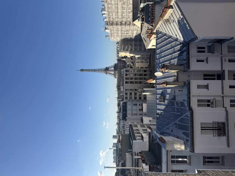 The image displays an elevated panoramic view of the city of Paris, France, under a clear, bright blue sky with minimal cloud cover. The Eiffel Tower is prominently positioned in the background, centered within the frame, rising above the urban sprawl. In the foreground and mid-ground, a dense collection of multi-story buildings, characteristic of Parisian architecture, features light-colored facades and a variety of rooftops. Many roofs are constructed from grey or silver-toned corrugated metal, while others appear tiled. Numerous chimneys and several white satellite dishes are visible on the rooftops. A large building in the mid-ground on the right is completely covered in white scaffolding, indicative of construction or renovation. In the far distance to the left, several modern high-rise buildings and an industrial structure emitting a white plume are discernible. No human figures or active interactions are visible. Shadows suggest direct sunlight.