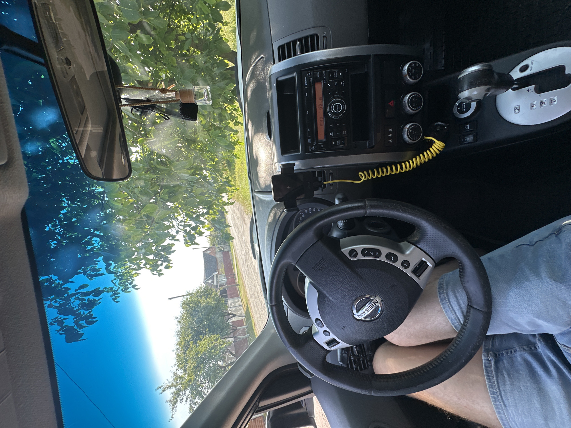 This is a first-person interior shot of a Nissan SUV parked on a dirt road in Shatava, Ukraine. The driver, a person wearing denim shorts, has their legs visible in the frame. The dashboard is in focus, showing the steering wheel, the center console with the radio and gear shift, and a phone mount with a charging cable. Outside the car, trees and houses are visible, indicating a rural setting. The sun is shining, casting shadows and creating lens flare on the windshield.
