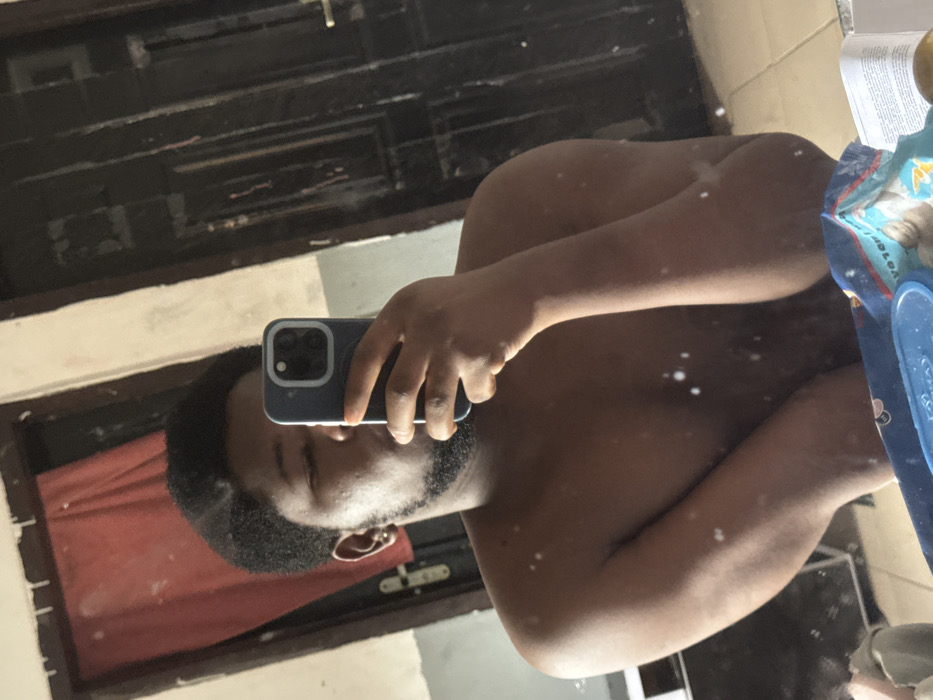 In an indoor setting within the city of Ipetumodu, Nigeria, a male individual with dark skin, short dark hair, and a beard is depicted in a mirror selfie. He appears shirtless, holding a dark-colored smartphone with his right hand, positioned to capture his reflection. A silver stud is visible in his left earlobe. His eyes are partially closed.
The reflected background features a light-colored wall section to the left, partially obscured by a hanging red fabric or curtain. To the right, a dark, paneled wooden door with a gold-colored doorknob is visible. The lower portion of the reflection shows a surface holding a blue plastic container, a blue and white packaged item (resembling wipes or diapers), a folded piece of paper with visible text, and a spherical object. Multiple small, white smudges are present on the mirror's surface, particularly across the reflection of the individual's torso. The environment exhibits signs of wear.