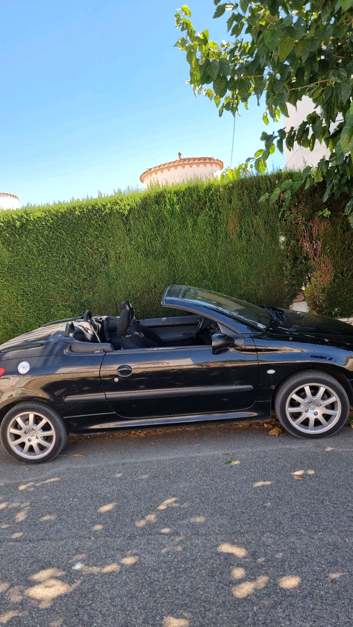 A black convertible Peugeot 206 CC is parked on the right side of a paved road in front of a manicured green hedge. The car is facing right and its top is down, revealing the black interior and seats. The hedge forms a barrier behind the car, and beyond the hedge, glimpses of two rounded structures with terracotta-colored roofs are visible against a clear blue sky. Sunlight casts dappled shadows from unseen foliage onto the road surface. The scene appears to be in a residential area, possibly in Castelló d'Empúries, Spain, given the architectural style of the roofing and the lush vegetation.