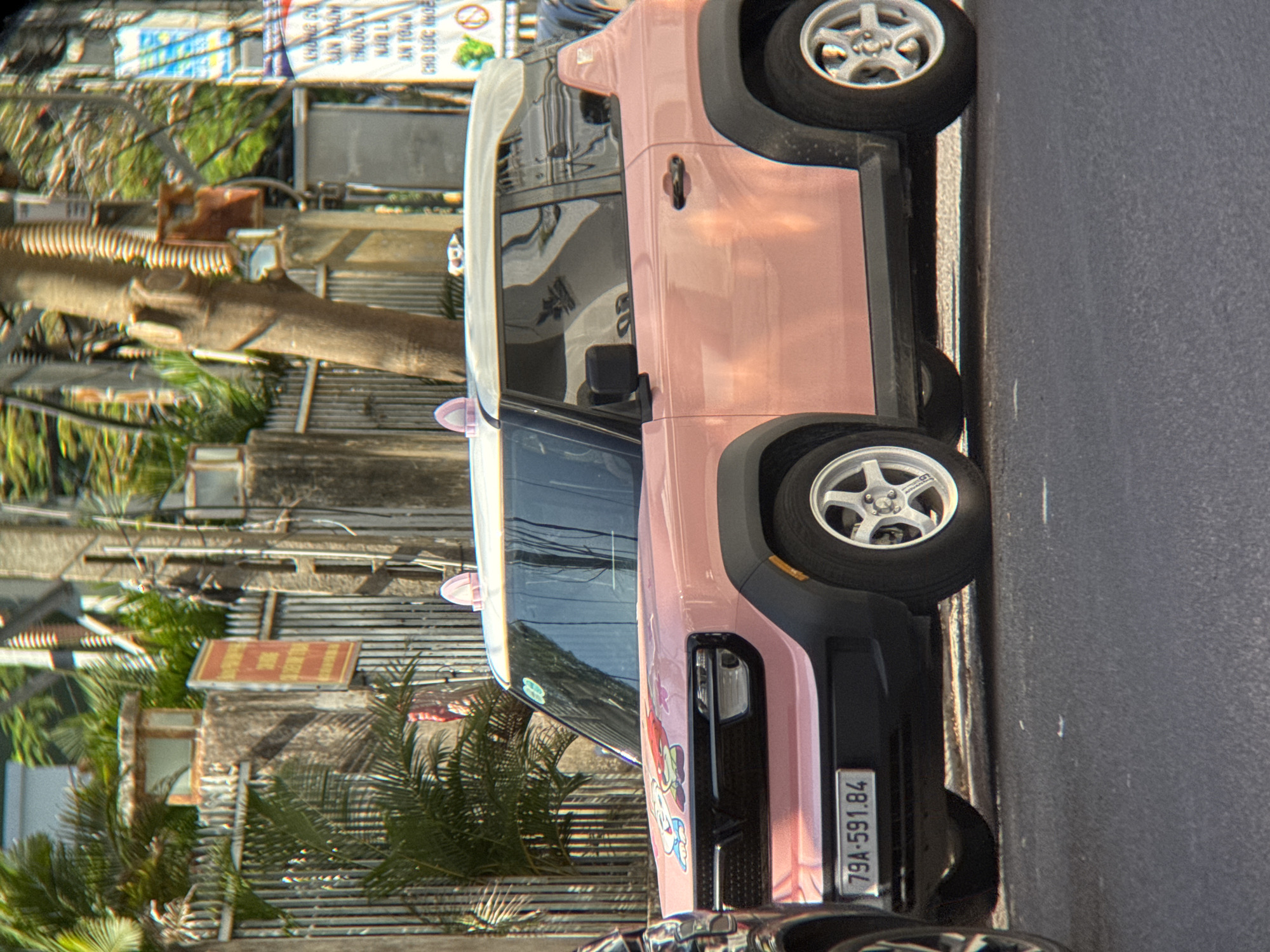 In Nha Trang, Vietnam, a light pink compact SUV, possibly a custom-painted Ford Bronco Sport, is parked on a paved street. The car has a white roof and two pink, cat-ear-shaped accessories attached to the top. The hood of the car is adorned with cartoon stickers, including a character resembling Hello Kitty and another with a red ribbon. The license plate reads "79A-591.84". The background features a concrete fence with a vertical railing and a red sign with Vietnamese text. Behind the fence, palm trees and foliage are visible, along with a utility pole and more signs. The overall scene suggests a typical streetscape in a Vietnamese city.