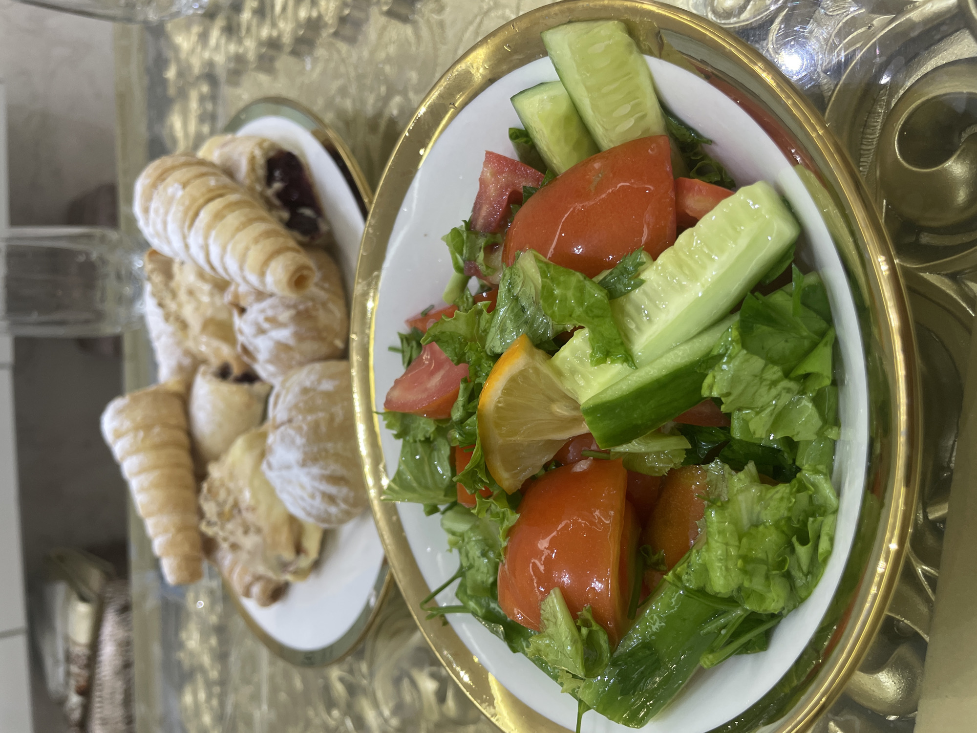 An overhead shot shows a salad and pastries. The salad, in the foreground, is in a white bowl with a gold rim, containing lettuce, chopped tomatoes, cucumber slices, and a lemon wedge. In the background, a plate of pastries, some dusted with powdered sugar, is visible. The setting appears to be a dining table in a home in Dushanbe, Tajikistan.