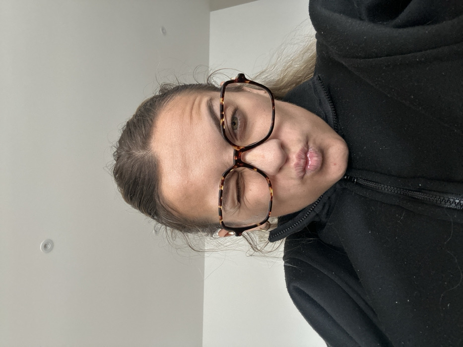 A single female individual is depicted in a close-up, mid-chest to head shot, angled slightly upwards. She has brown hair, pulled back, with visible wrinkles on her forehead. She is wearing large, square-shaped eyeglasses with tortoise-shell frames. Her right eye (viewer's left) is open, looking directly forward, while her left eye (viewer's right) is closed in a wink. Her lips are pursed into a forward, puckered expression. She wears a black, high-collared garment with a visible zipper running down the front. The background is a plain, light-colored ceiling, with a single circular recessed light fixture positioned in the upper center of the frame. The scene is indoors, in Veauchette, France.