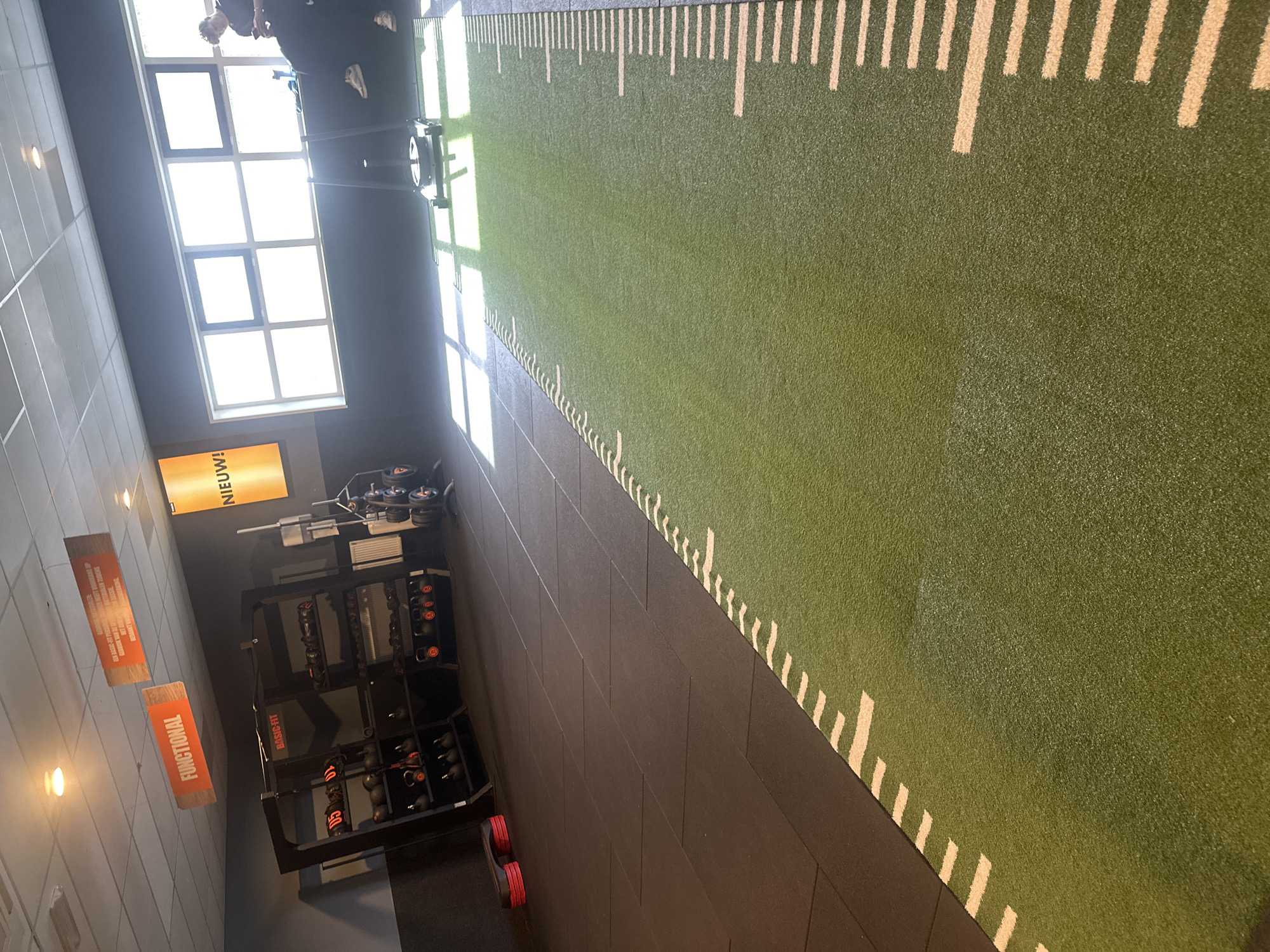 This image shows the interior of a Basic-Fit gym, likely located in the city of Schagen, Netherlands. The view is from a high angle, capturing an array of fitness equipment, including dumbbell racks loaded with weights, a functional training area, and an artificial turf track with markings that appear to be measurement lines. Bright daylight is filtering in through a set of large windows, casting strong shadows across the room. Various signs are visible, including a Basic-Fit logo, indications for the functional area, and a sign written in Dutch. The gym floor is a combination of dark tiles and green turf, creating designated spaces for different types of workouts. The environment seems well-lit and organized, with equipment neatly arranged.