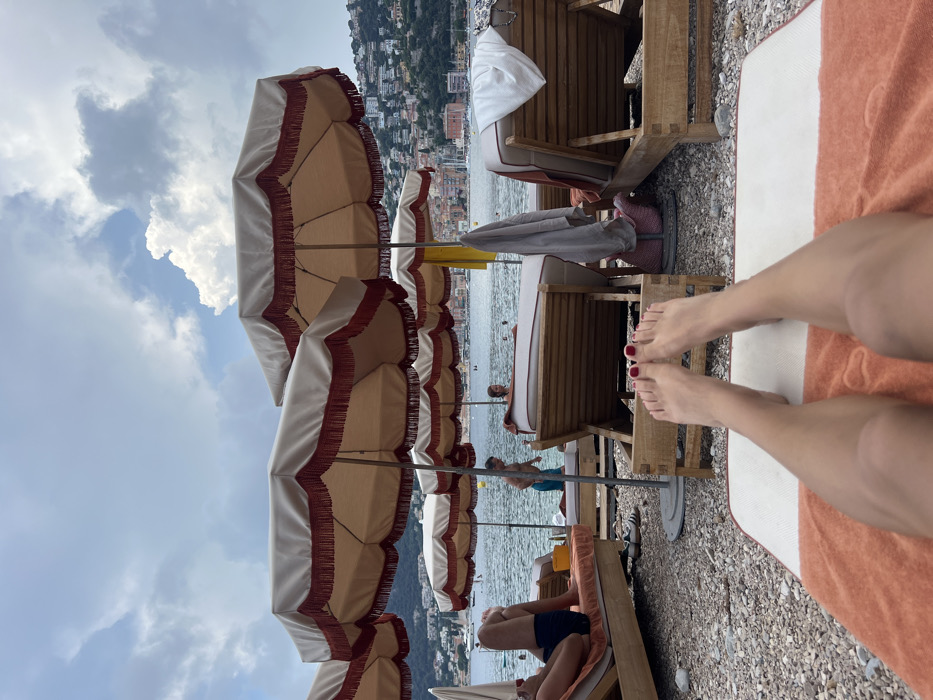 A sunny beach scene features rows of beach umbrellas with white canopies and red fringe. Lounge chairs sit beneath the umbrellas, with people relaxing on them. In the foreground, a person's legs are stretched out on a beach towel. Their toes are painted red. The beach is pebble-strewn and the water is visible in the background, with buildings climbing up a hillside. The sky is cloudy.