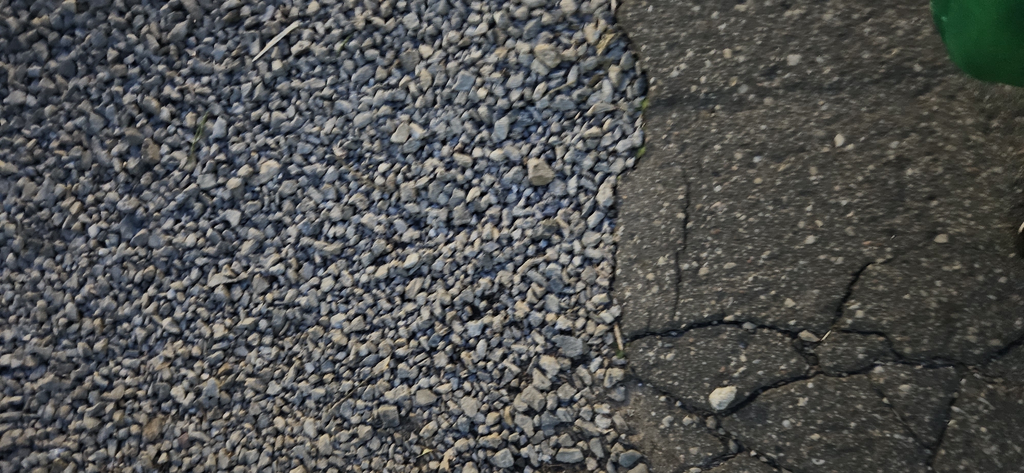 A close-up, high-angle shot captures a boundary between a gravel surface and cracked asphalt. The gravel, predominantly gray with some lighter-colored stones, fills the upper portion of the frame. The asphalt, dark gray with white speckles, occupies the lower portion, showing distinct cracks and a slightly uneven texture. A hint of green is visible in the bottom right corner, likely part of an object outside the main focus. The scene appears to be an outdoor urban environment. Without further context or visual cues, pinpointing the exact location within Vitsebsk, Belarus is not possible.