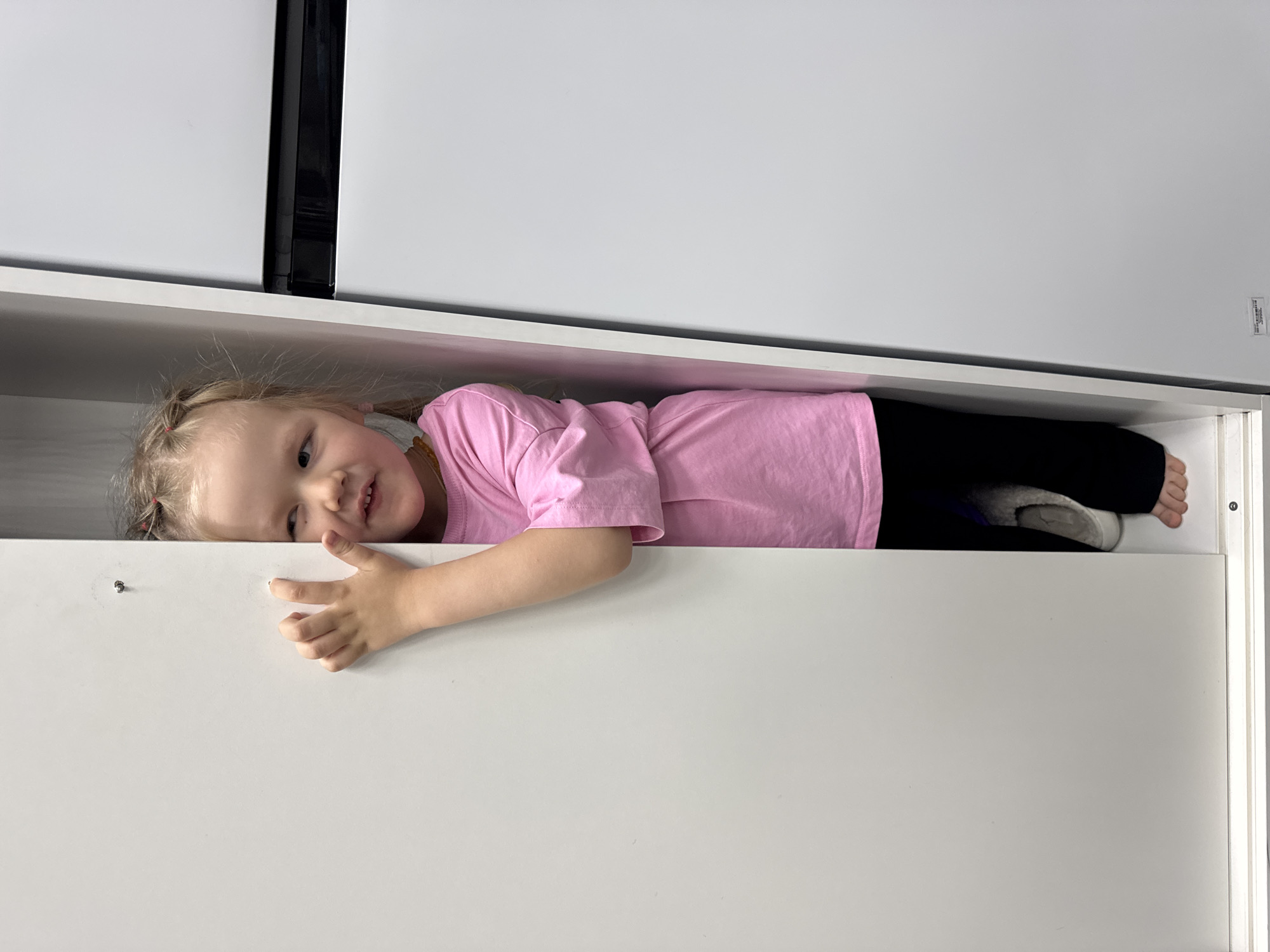 A young child wearing a pink shirt and black pants is lying inside an open white drawer. The child has blonde hair styled in pigtails. The drawer appears to be part of a white cabinet or storage unit. The location is unspecified and cannot be determined. The exact location in the city of Perm, Russia is not present within the image.
