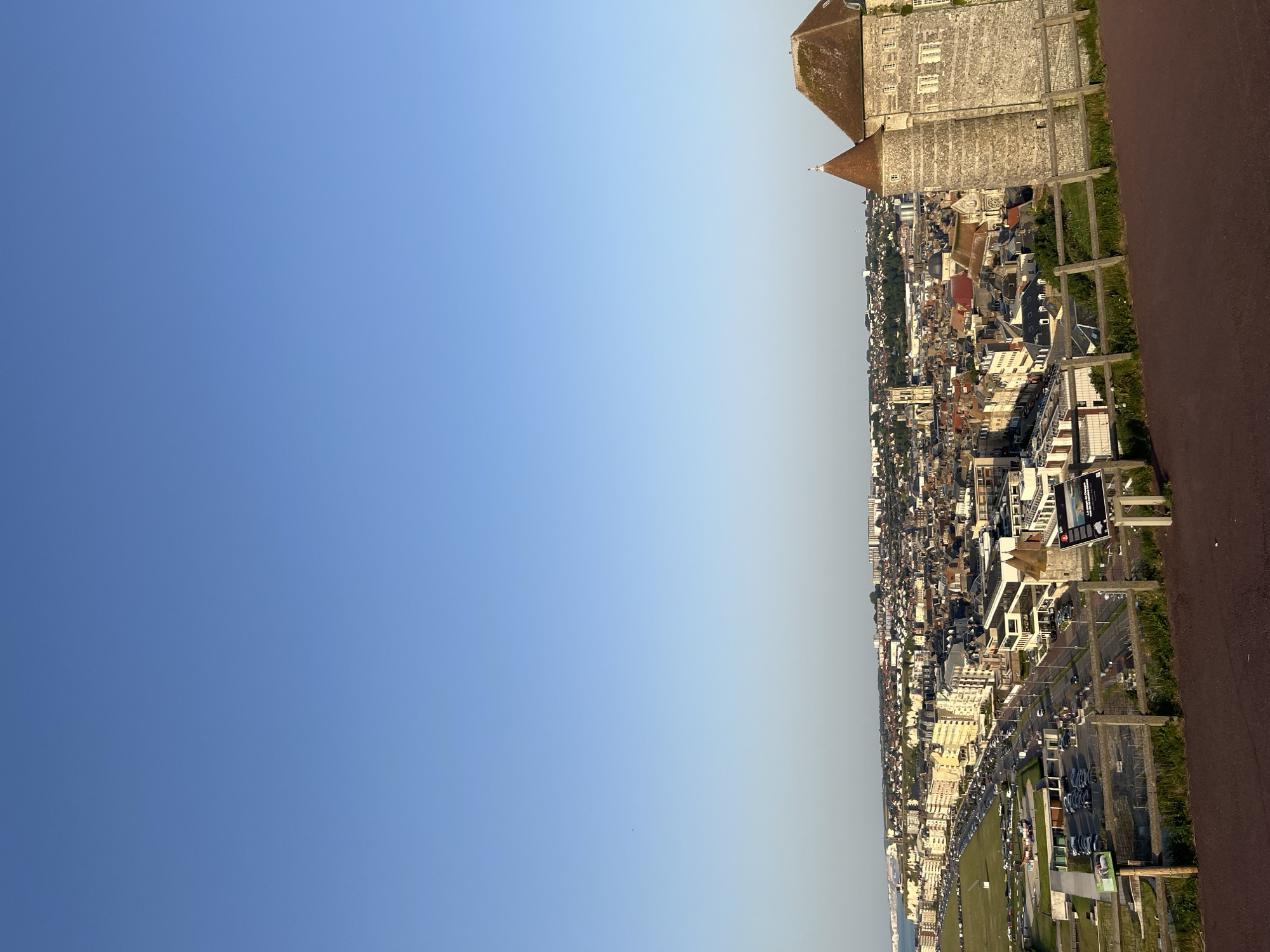 This is a landscape photograph taken in Dieppe, France, from an elevated viewpoint looking down on the town and its coastline. The image is oriented vertically, with the sky occupying the left two-thirds and the town and coastline on the right. The sky is a clear, bright blue with a slight gradient towards a lighter hue near the horizon.
The town of Dieppe stretches along the coast, characterized by a dense collection of buildings with red-tiled roofs, typical of European architecture. Several modern buildings with white facades are interspersed among the older structures. A prominent stone building with a conical roof, possibly a historical landmark or fortification, is visible at the top right of the image, overlooking the town. Below it, a church with a dome is visible.
Along the coast, a street lined with parked cars and some grassy areas is present. There are also several large billboards visible in this area. A hint of the sea or coastline is visible in the distance on the far right, suggesting the town's proximity to the water. The overall scene depicts a coastal town on a clear day, likely in the late afternoon or early evening, given the angle of the sunlight casting light and shadows. There are no specific activities or events clearly depicted, but the presence of cars suggests it is a populated area. The overall emotion conveyed is one of a serene, sunny day in a historic French coastal town. Visible text includes that on the billboards, but it is not legible.