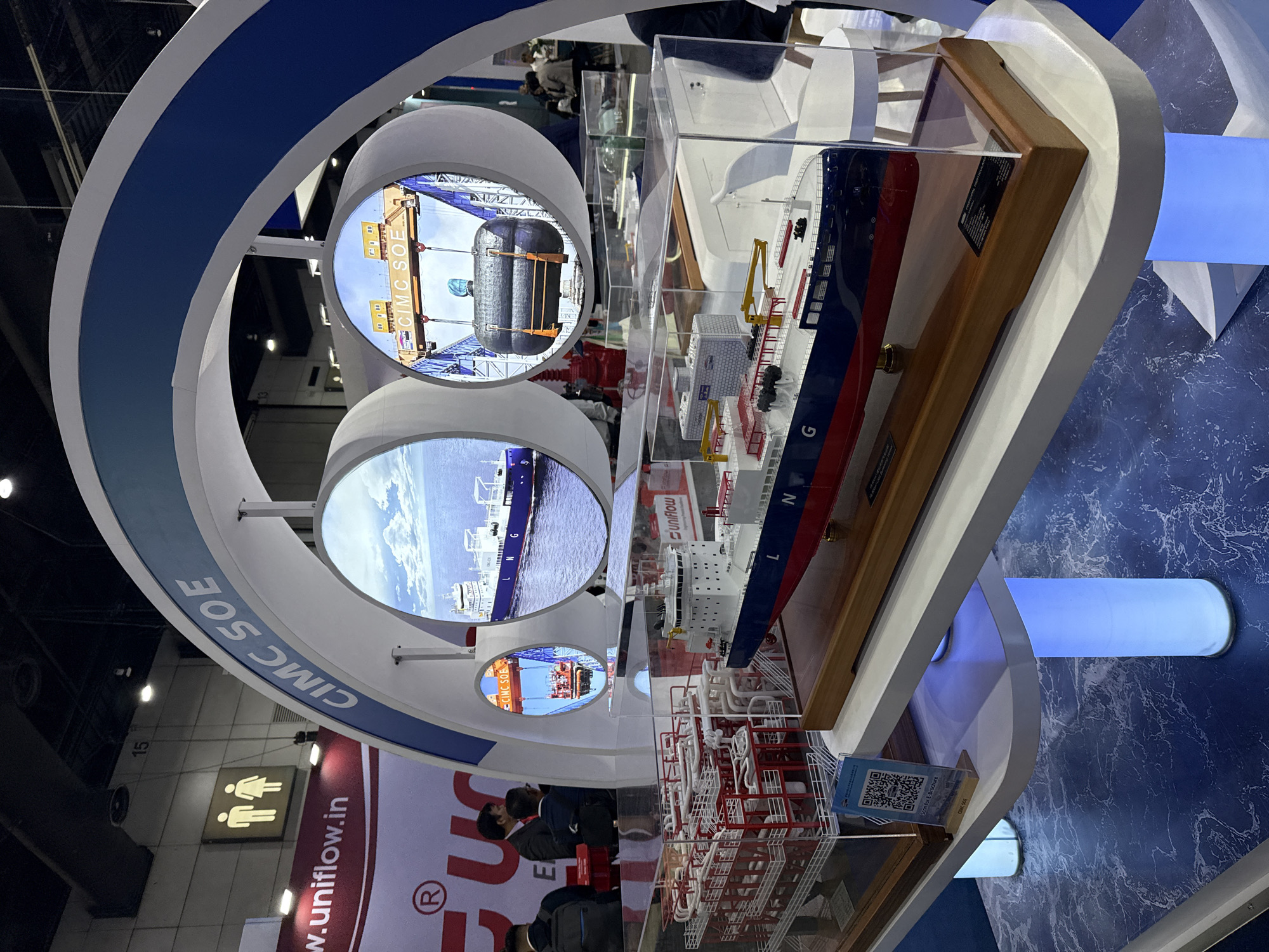 The image shows a display at an exhibition, likely a trade show, featuring models of ships and industrial equipment. In the foreground, a detailed model of an LNG carrier ship is prominently displayed on a white shelf, with a clear acrylic case protecting it. To the left of the ship model is a smaller model of a complex industrial facility with red and white structures and intricate piping. The entire display is set against a backdrop that includes circular screens showing images related to shipping and maritime operations, with the text "CIMC SOE" visible on the structure above. The background shows a trade show environment with other booths, signage, and people walking around. The floor beneath the display is patterned to resemble water. The presence of a QR code with the text "Scan for E-Brochure" suggests promotional material is available. The scene is brightly lit, indicating an indoor event space. There is no specific indication of the city being Bangkok, Thailand, within the image itself.