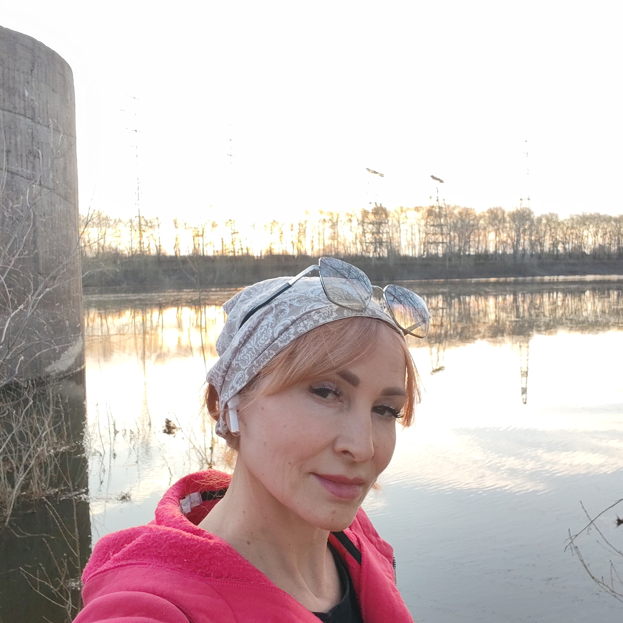 A woman in a pink hooded jacket and paisley-patterned headscarf stands in front of a body of water. Sunglasses are perched on top of her headscarf. She has light-colored hair and wears an earpiece. In the background is a concrete structure, possibly a support for a bridge or dam. Across the water, there are trees and utility poles visible, reflecting in the water. The location is likely the city of Chaikovsky, Russia, based on the concrete structure and landscape, though specific landmarks would be needed for verification. The lighting suggests it is either sunrise or sunset. The woman is looking directly at the camera, appearing to take a selfie.