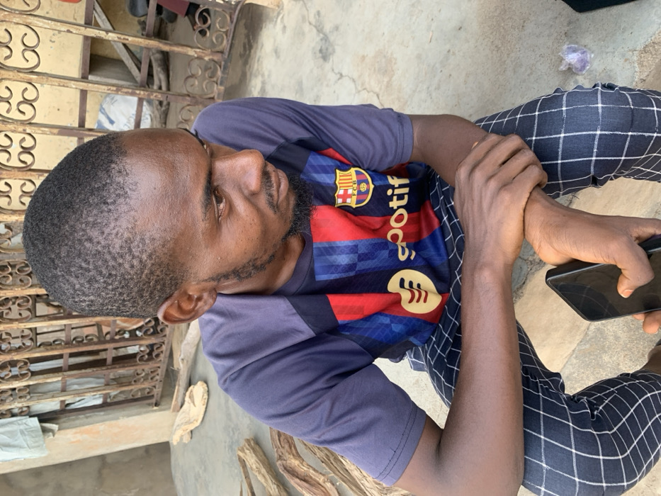 A young adult male, appearing to be in his late twenties to early thirties, with dark skin, short dark hair, and a short beard and mustache, is seated on a light-colored concrete surface. He is wearing a short-sleeved dark blue and striped red/blue sports jersey featuring the Spotify logo and FC Barcelona crest, along with dark-colored trousers displaying a white grid pattern. His gaze is directed downwards and to his right. His left hand holds a black smartphone, while his right hand rests on his left forearm. Behind him, a light-colored building wall is visible, along with a dark brown or black ornate metal gate or grille. Several pieces of dried, light-colored wood are scattered on the ground to his left, and a small crumpled piece of plastic or paper is on the ground to his right. The scene is located in Okene, Nigeria.