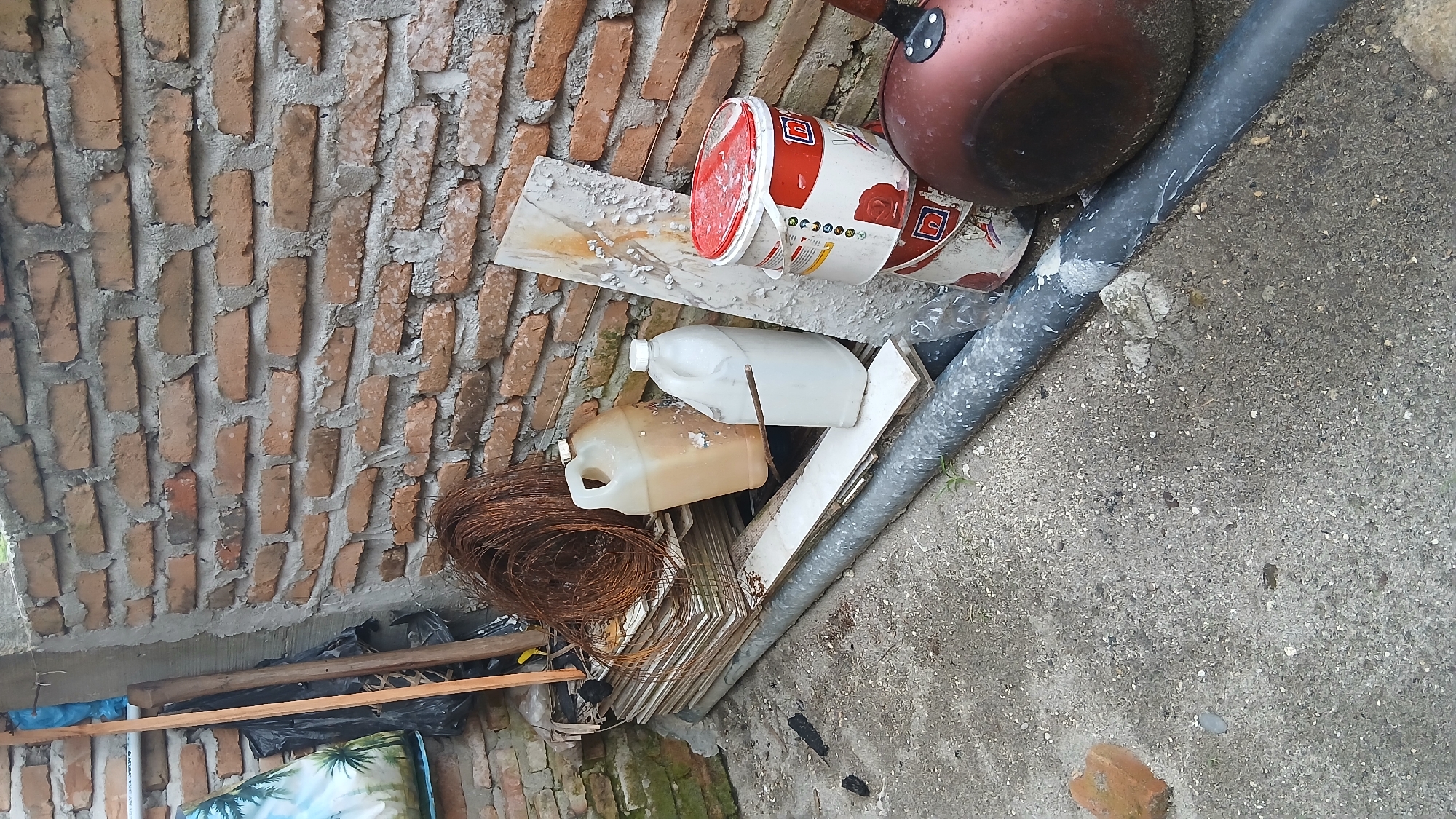 The image depicts a cluttered outdoor scene against a brick wall. In the foreground, a grey pipe runs across the concrete ground. To the left, a pile of flat, light-colored materials is stacked. On top of this stack, a large coil of rusty wire is visible. Next to the wire, there are two plastic containers: a larger, tan-colored one and a smaller, white one. Further to the right, leaning against the brick wall, is a long, thin piece of material with a textured, peeling surface. Stacked on the ground next to this are two large cylindrical containers, one white with a red lid and a red design, and another similar container below it. Partially visible to the far right is a dark red, round object, possibly a pot or pan. Behind the stacked materials and containers, a weathered brick wall forms the background. The ground is covered in concrete and small debris. The scene appears to be in a storage or work area. The location is identified as Klambir Lima Kebun, Indonesia.