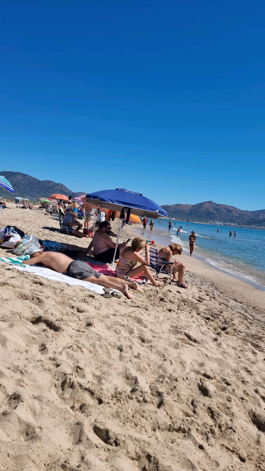 This image depicts a sunny day at a crowded beach in Castelló d'Empúries, Spain. The foreground is dominated by the sandy beach, with footprints and the texture of the sand visible. In the middle ground, several people are relaxing under umbrellas. A man is lying on a towel, facing away from the camera, wearing grey shorts. Next to him, a man and a woman are sitting in chairs under a blue umbrella. The woman is wearing a patterned bikini. Further back, more people are visible, some sitting under umbrellas and others walking towards or in the shallow water. Several colorful beach umbrellas are scattered across the sand. The background features the blue Mediterranean Sea with gentle waves and a distant coastline with hills and buildings. The sky is clear and bright blue.
