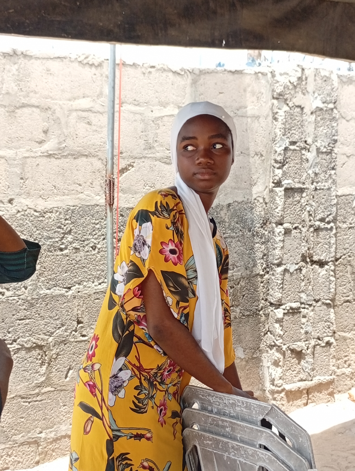 A young woman wearing a yellow floral dress and a white headscarf stands next to a textured, concrete-block wall. She is holding a stack of grey, metal chairs. An unidentified arm is partially visible on the left side of the image. The location is Adankolo, Nigeria.