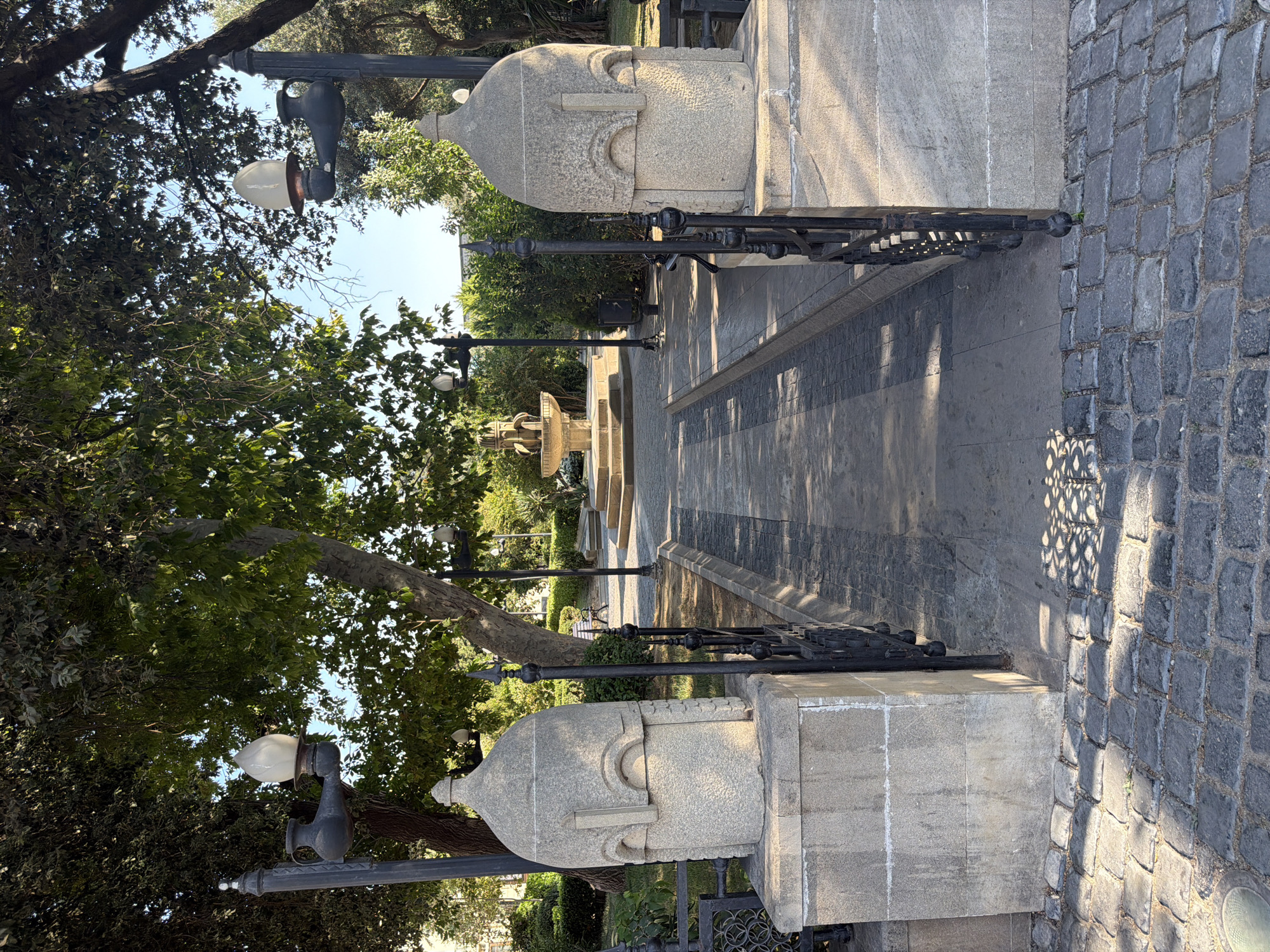 The image depicts a park entrance in Sabail Raion, Azerbaijan. Two large, intricately carved stone pillars flank a pathway leading into the park. Each pillar is topped with a stone structure resembling a helmet or a stylized head, and a lamp post with a pitcher-shaped fixture and a bulbous glass shade rises behind each pillar. A black wrought-iron fence runs between the pillars and along the edge of the pathway. The pathway is paved with dark grey and lighter grey rectangular stones, creating a striped pattern. Beyond the pathway, a fountain with multiple tiers is visible in the distance, surrounded by lush green trees and manicured bushes. The ground in the foreground is paved with dark cobblestones, and sunlight casts dappled shadows on the pathway and ground. A few distant figures are walking in the park. The environment appears to be an urban park on a clear day.