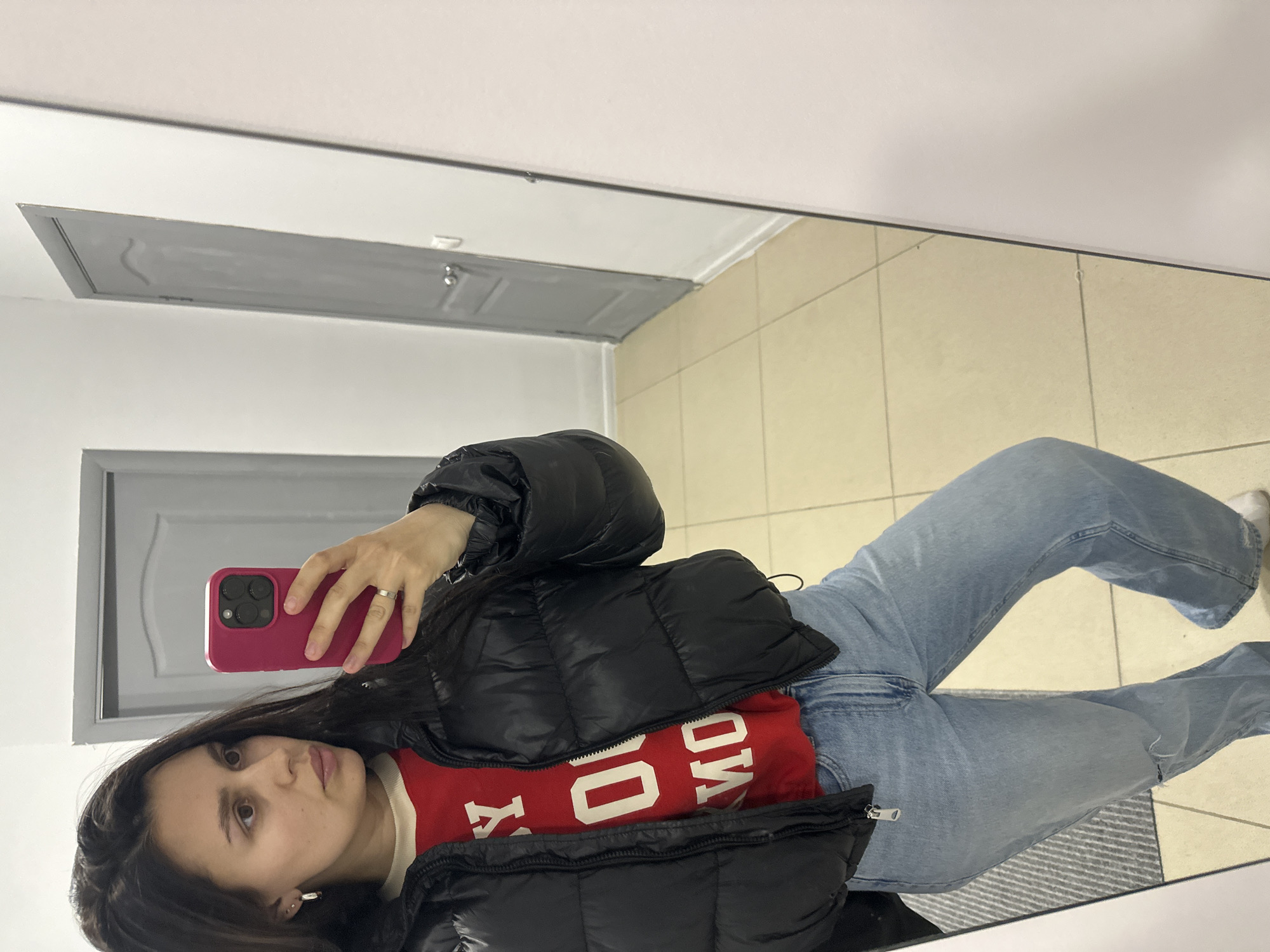 A young woman is captured in a mirror selfie, standing indoors in what appears to be a room or hallway in Chaikovsky, Russia. She holds a vibrant pink-red smartphone with a dark camera module in her right hand, positioning it to reflect her upper body and legs. Her long, dark hair frames her face, and she has a neutral, observant expression. She wears a silver ring on her left ring finger and small earrings.
She is dressed in a black, shiny puffer jacket, worn open to reveal a red long-sleeved top underneath. The red top features white lettering, which, when read as a reflection, clearly spells "TOMMY" or a similar brand name. She also wears light blue, wide-leg jeans, with white socks visible on her left foot.
The background, seen in the mirror's reflection, consists of plain white walls and a light-colored tiled floor, possibly beige or cream, with visible grout lines. To the left, a light gray door with a recessed panel design and a silver doorknob is visible. The ceiling is white, reflecting a long, rectangular light fixture or vent. A gray patterned rug is partially visible on the floor beneath her legs. The lighting is bright and even, suggesting artificial illumination.
